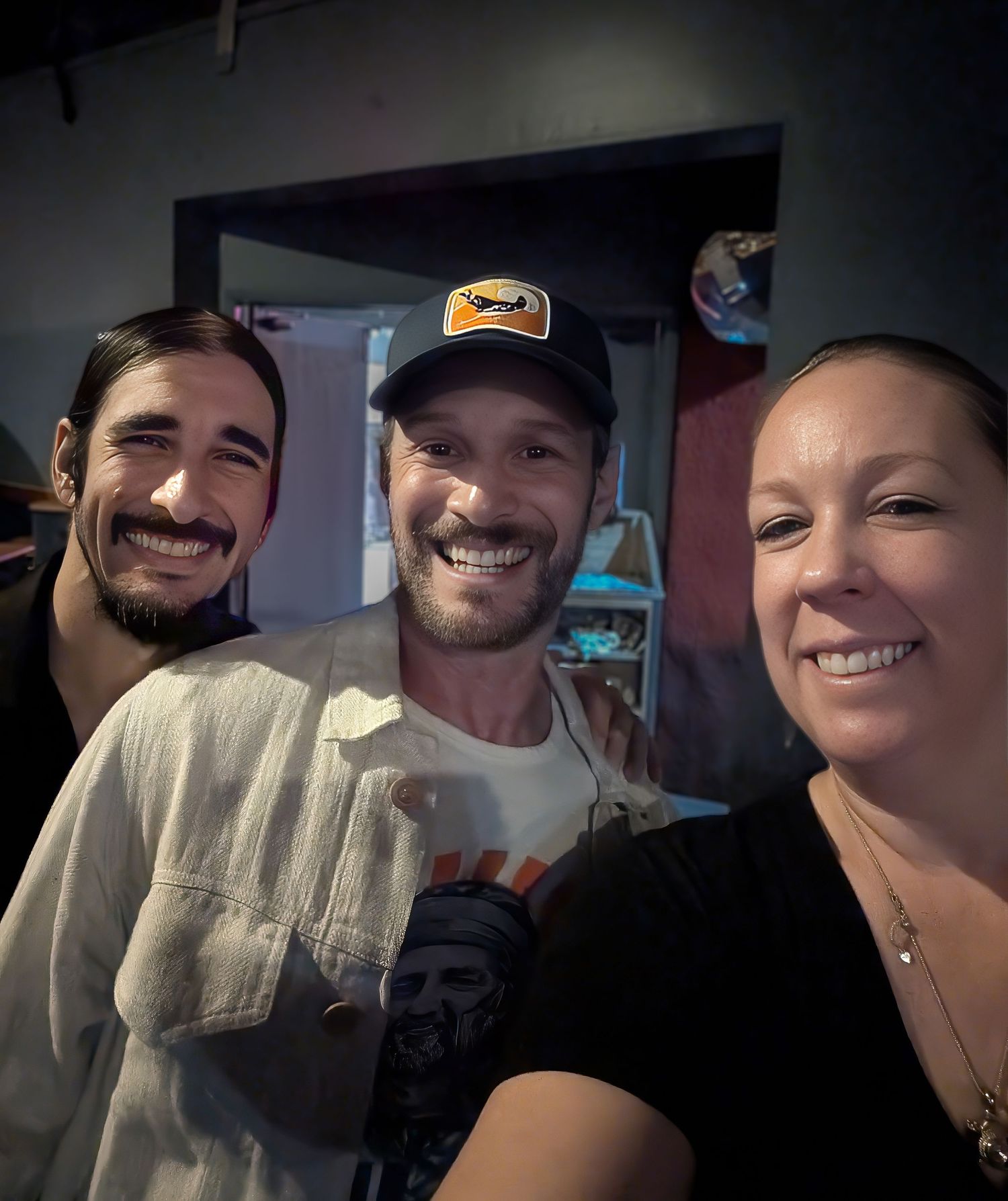 Josh Wolf comedian. Three smiling friends posing together for a selfie indoors, one wearing a baseball cap and graphic tee.