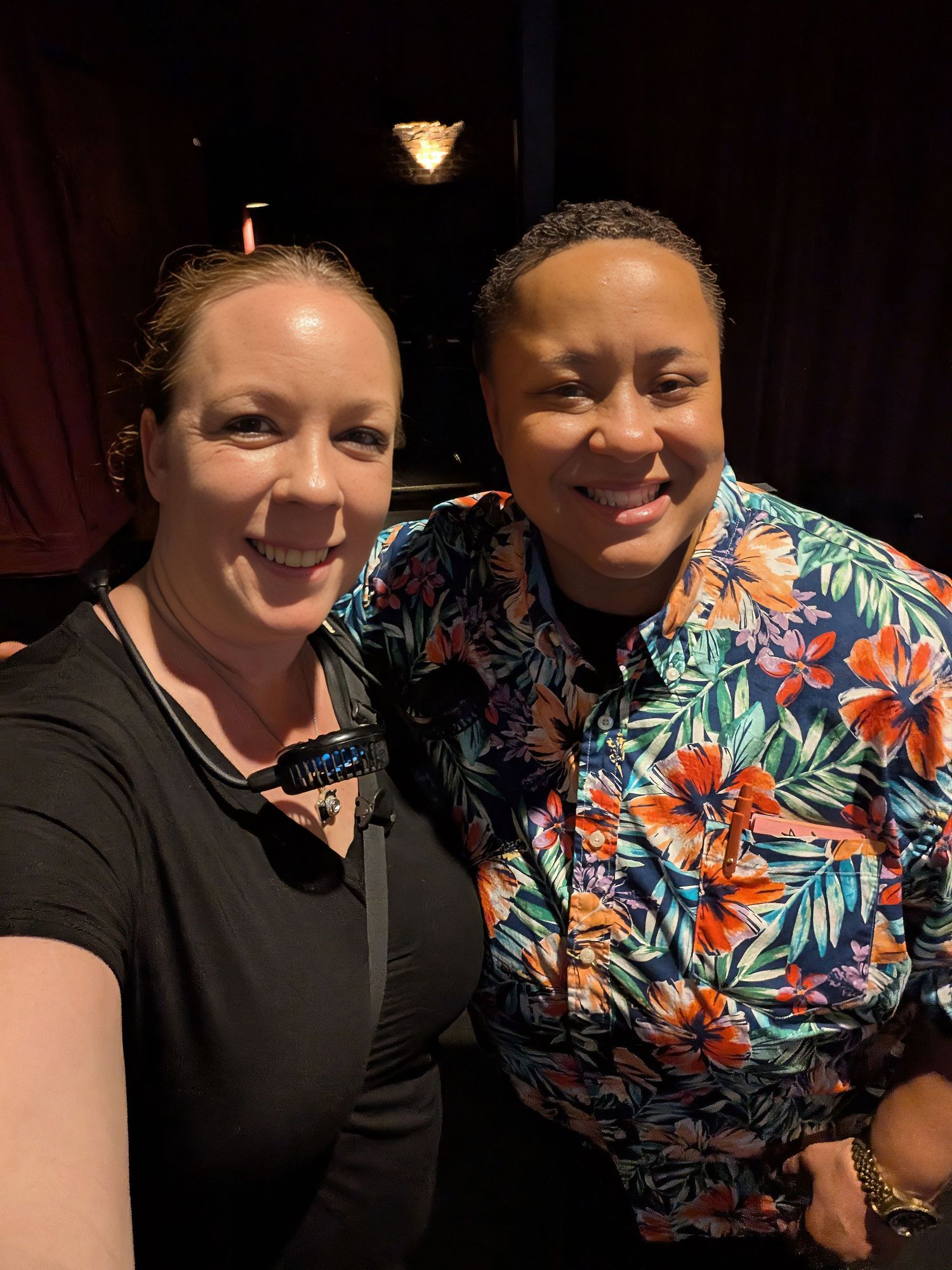 Nicole Starnes comedian.Two smiling women pose together for a selfie indoors, one wearing a black top and the other a colorful floral shirt.