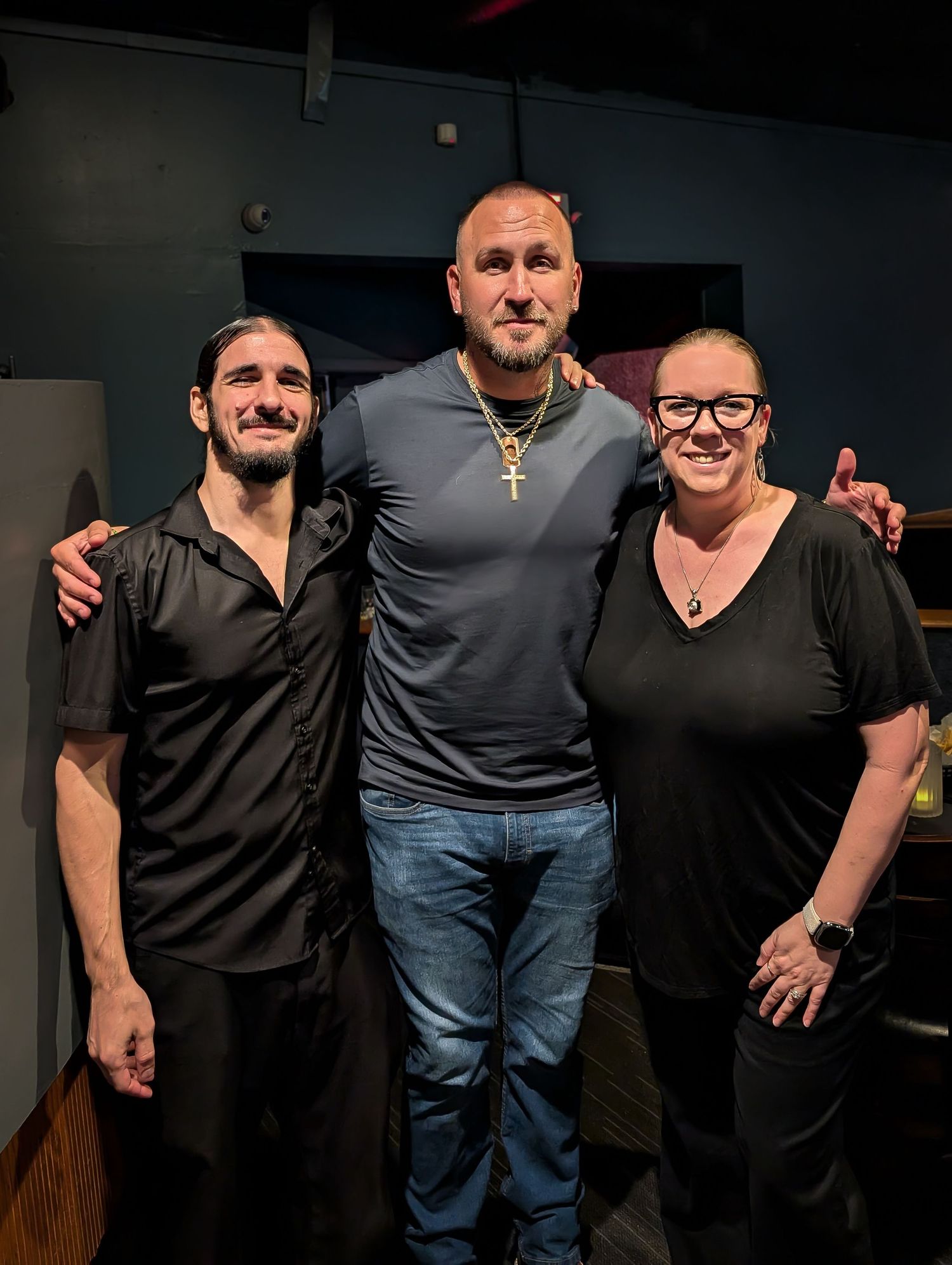 Kevin Eudy comedian. Three people smile together for a photo indoors, with a tall man in gray flanked by two friends in black.