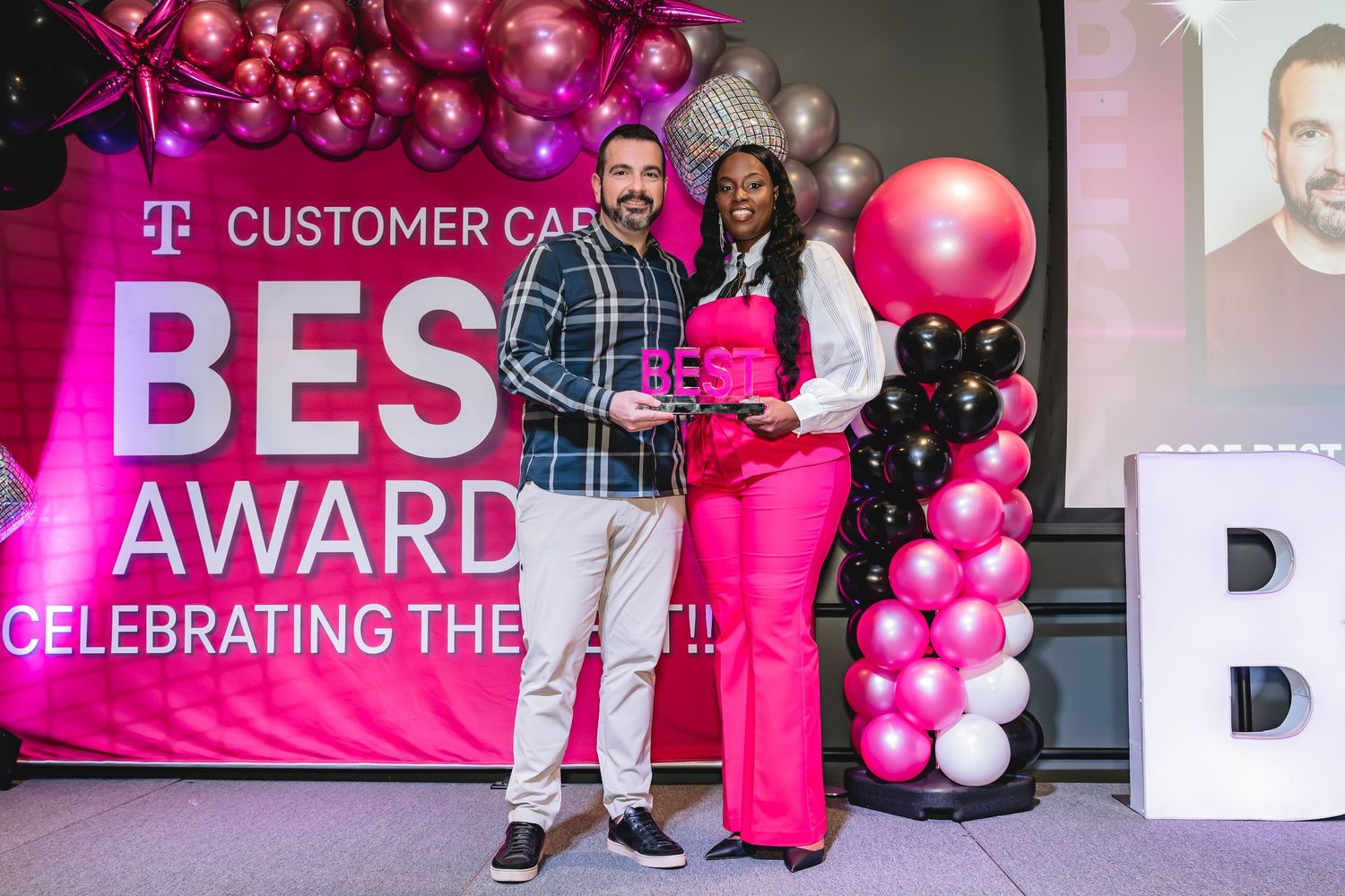 Two people pose with a pink award at T-Mobile Customer Care Best Award ceremony with balloon decorations.