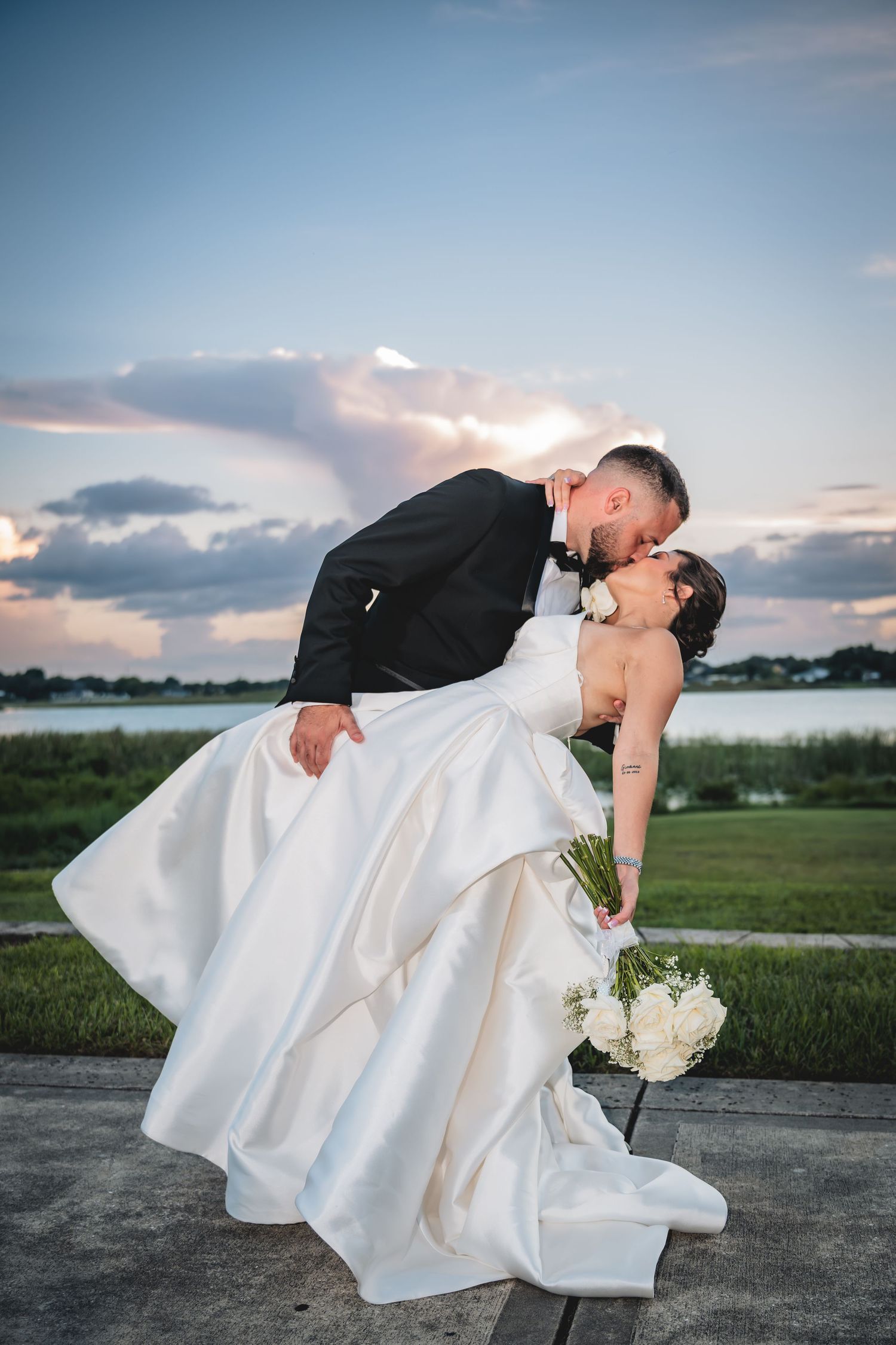 Groom dips bride for a romantic kiss at sunset near a waterfront, bride holding white bouquet in elegant white gown. Tampa Photographer