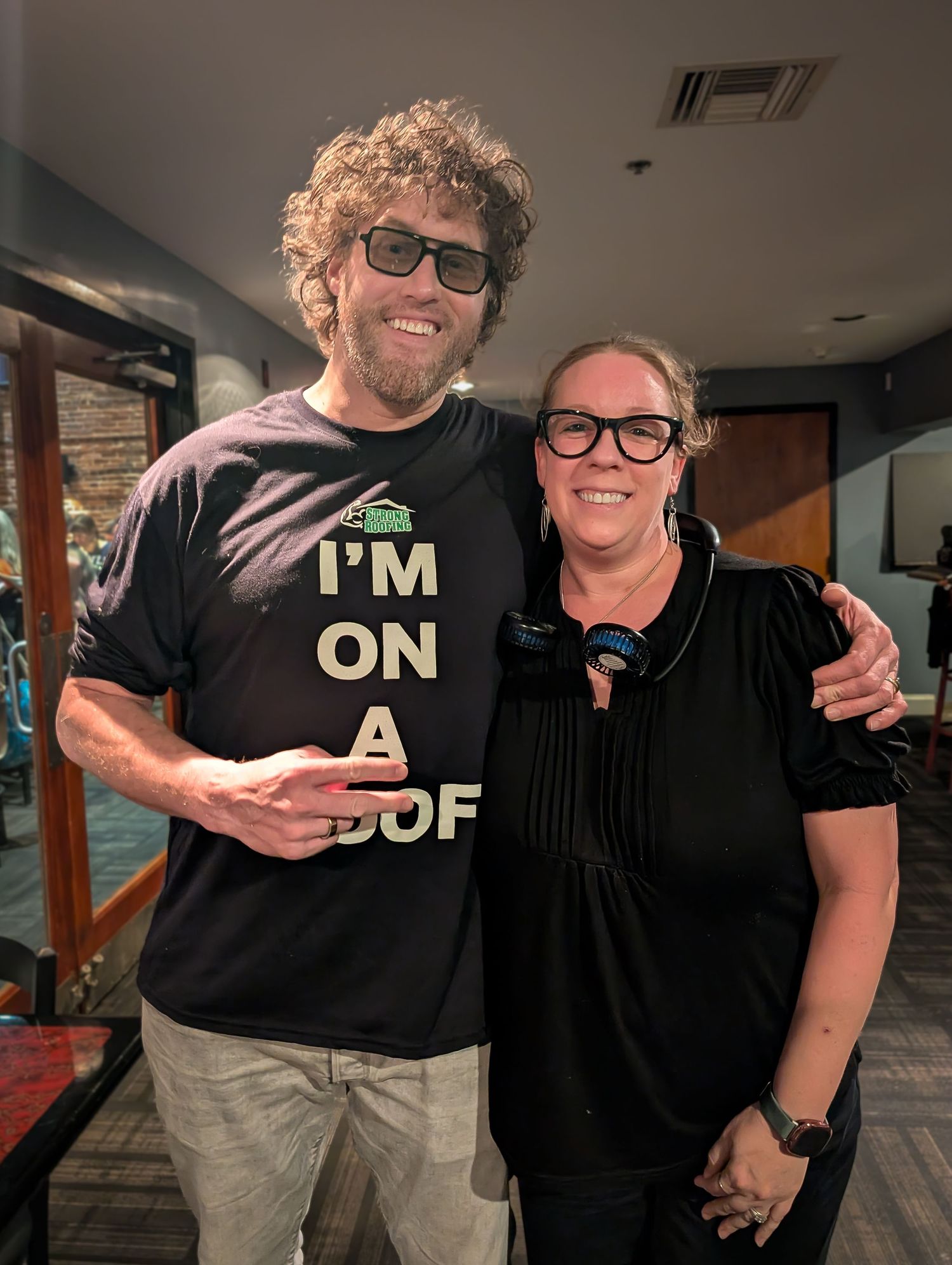 TJ Miller comediam and actor.Two smiling people pose together indoors, man wearing 3D glasses and I'm On A Roof t-shirt, woman in black top.