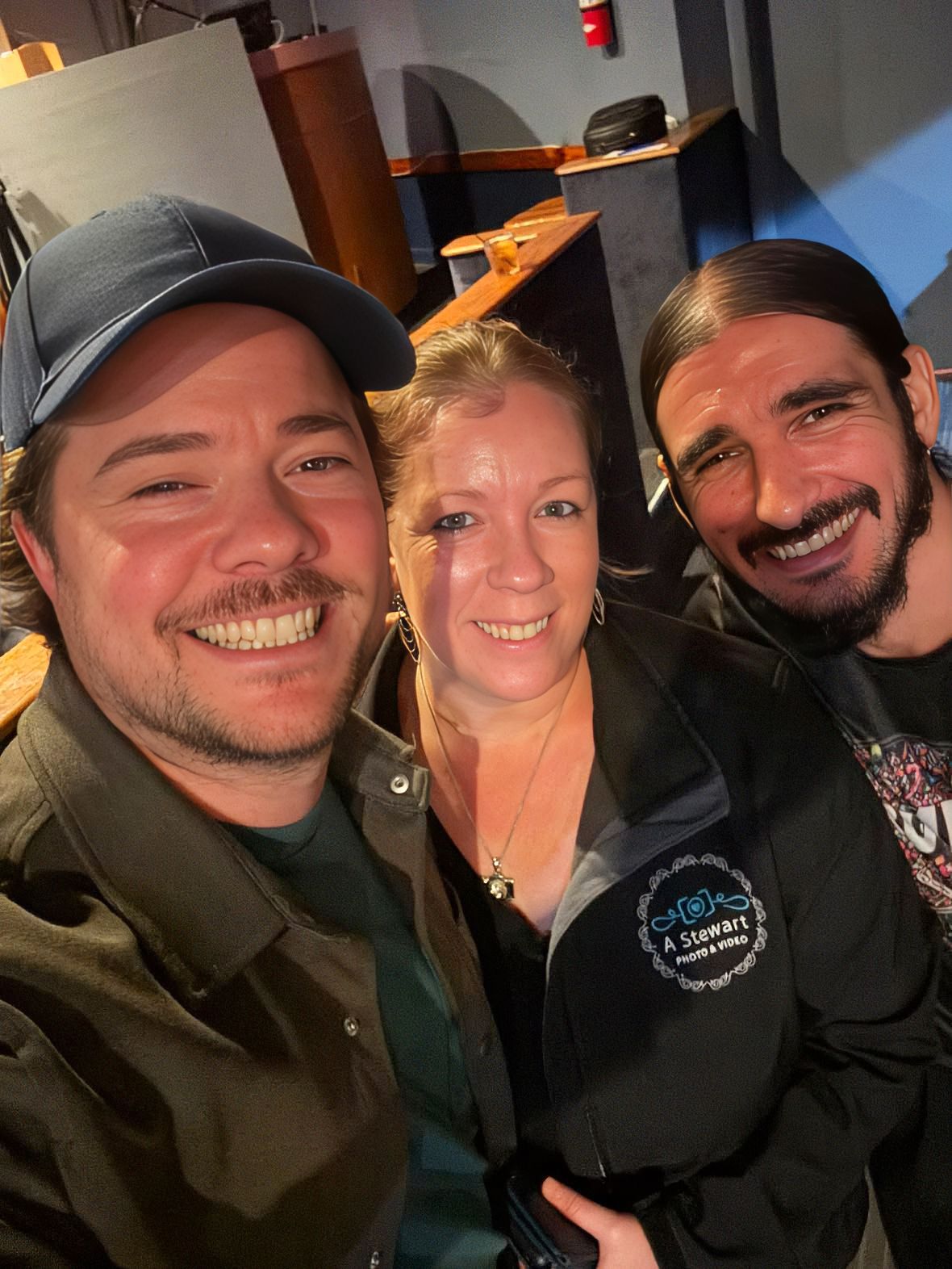 Zach MAMA comedian. Three smiling friends pose for a selfie indoors, with one wearing a blue cap and another in a branded jacket.