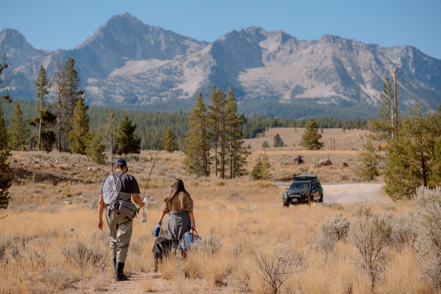A group of hikers traverse a scenic mountain trail with rugged peaks and evergreen trees in the background.