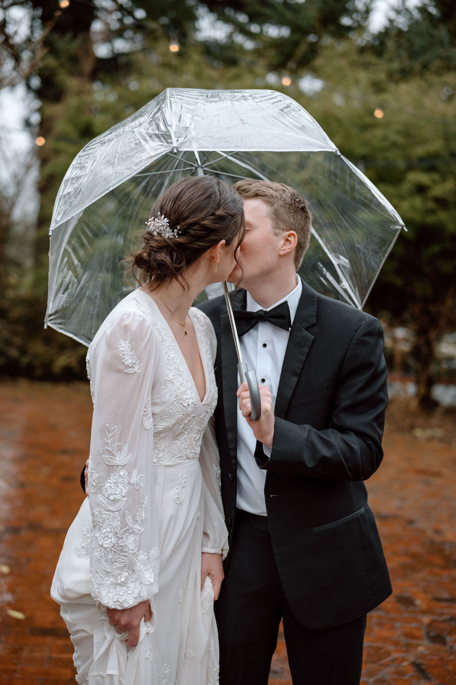 Newlywed couple shares intimate moment under clear umbrella in autumn garden with twinkling lights overhead.