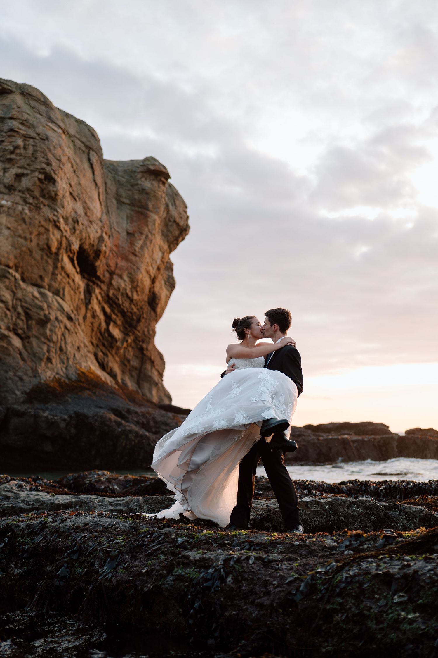 A romantic couple in formal attire embrace on rocky coastal cliffs at sunset with dramatic ocean views.