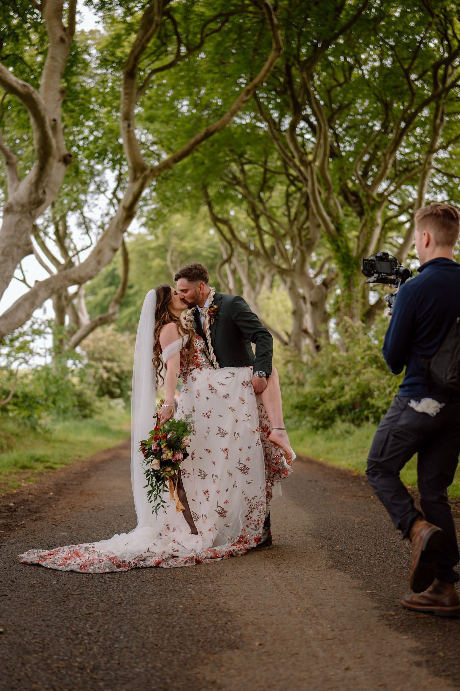 A photographer captures a romantic moment on a tree-lined path during a wedding photoshoot.