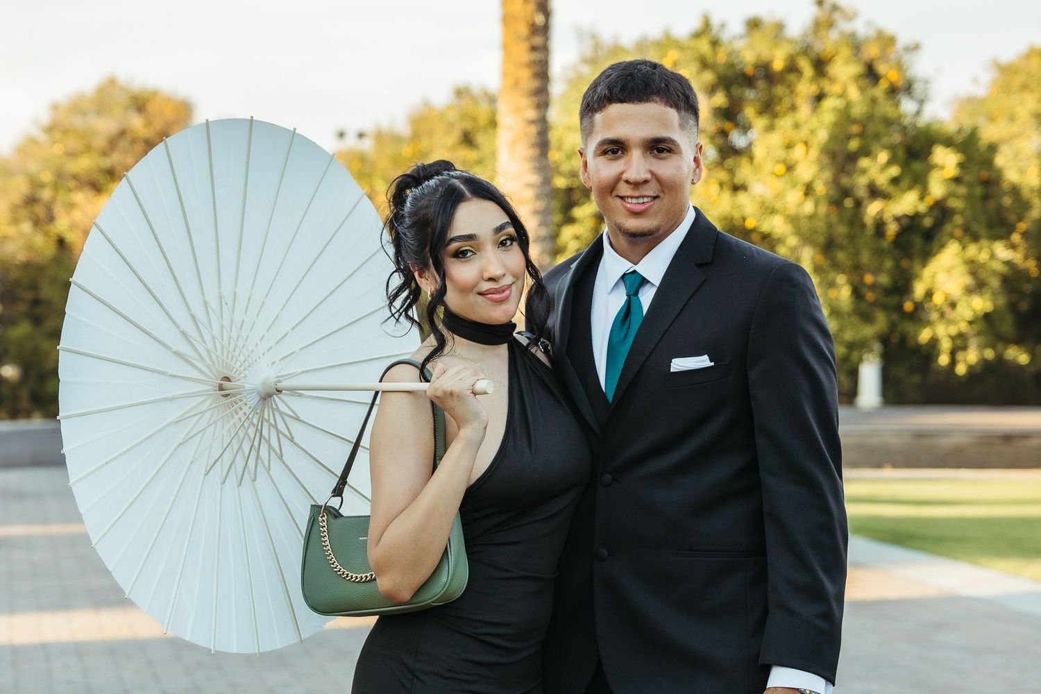 Couple in formal attire poses together outdoors with a white parasol at an event.
