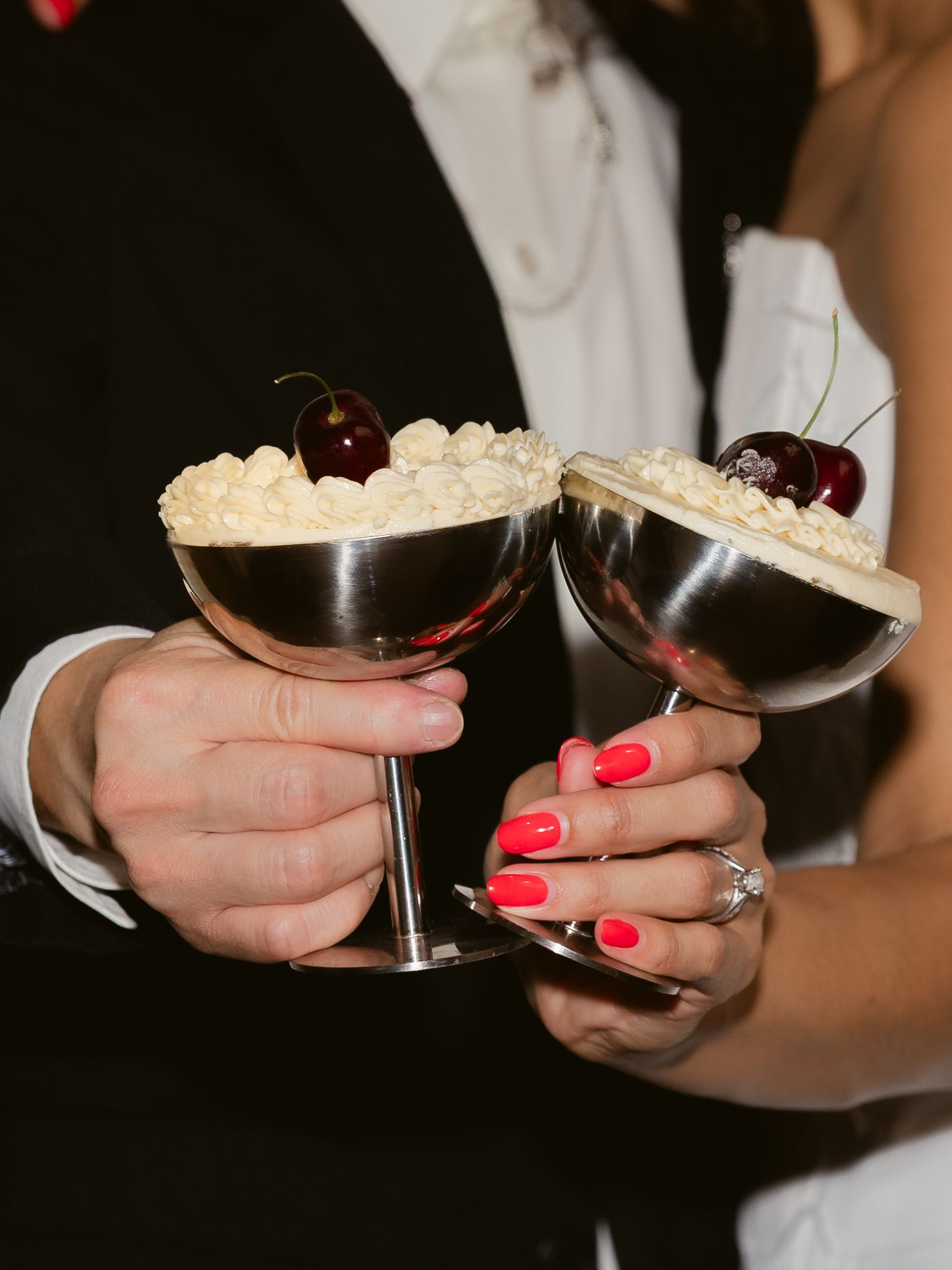 Bride and groom toasting with silver coupe cocktail glasses topped with whipped cream and cherries at wedding reception.