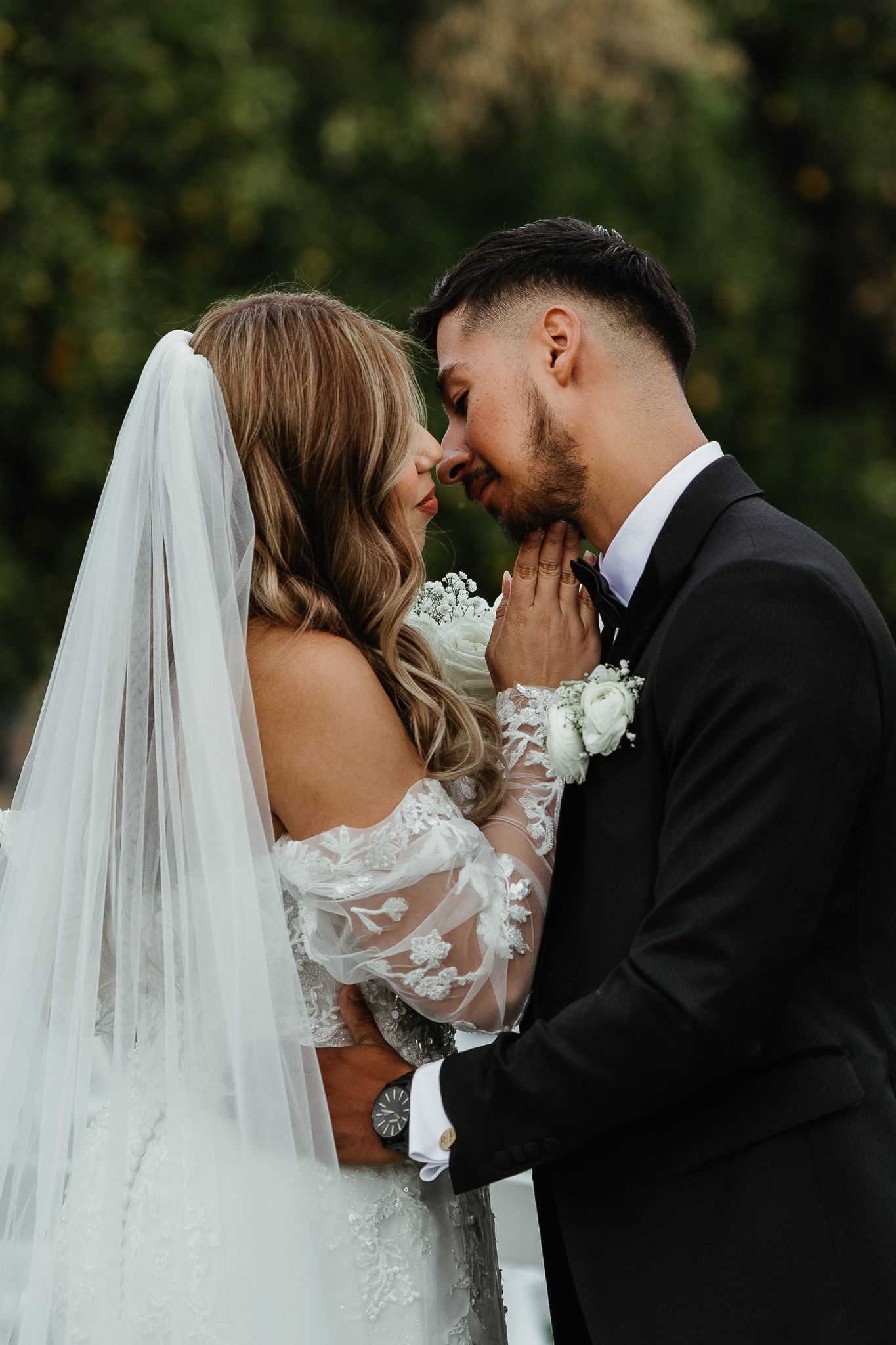 Bride in off-shoulder lace gown and veil touching groom's face as they lean in for a kiss outdoors.
