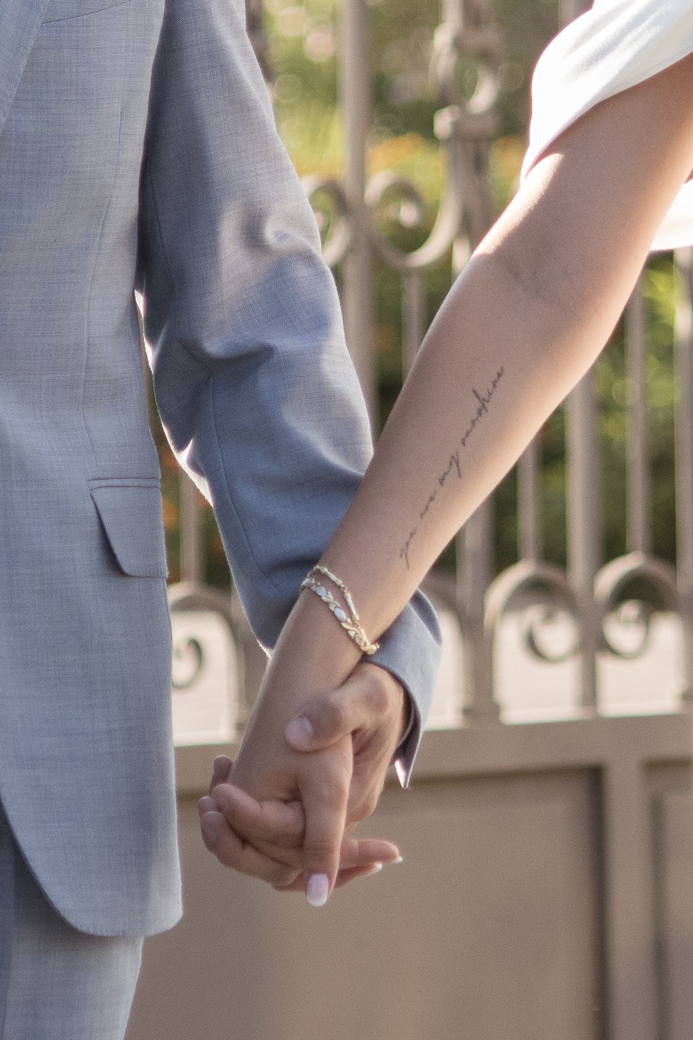 Bride and groom holding hands, bride wearing white gown with script forearm tattoo and gold bracelet.