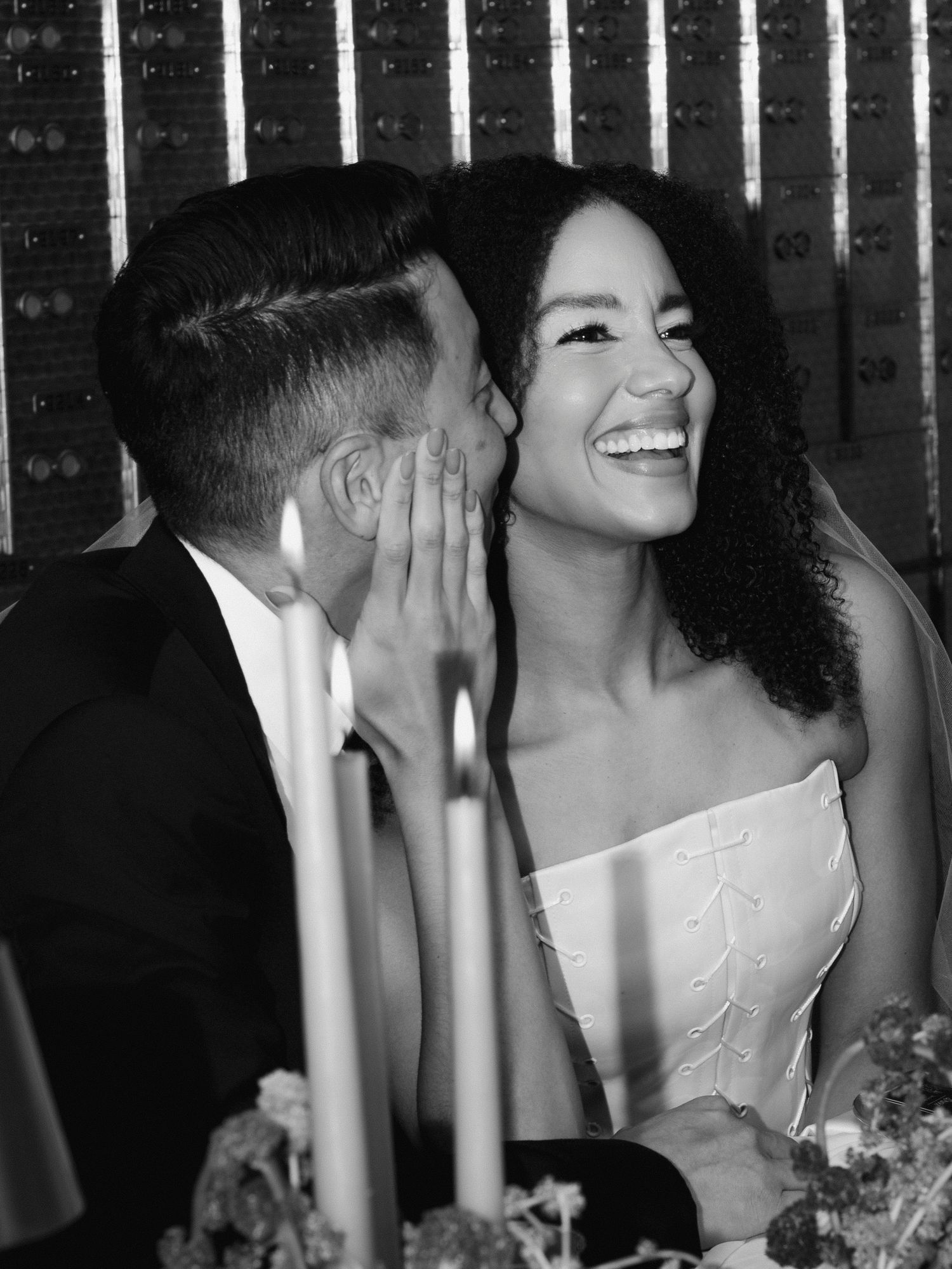 Black and white photo of bride laughing joyfully as groom whispers in her ear at wedding reception candlelit table.