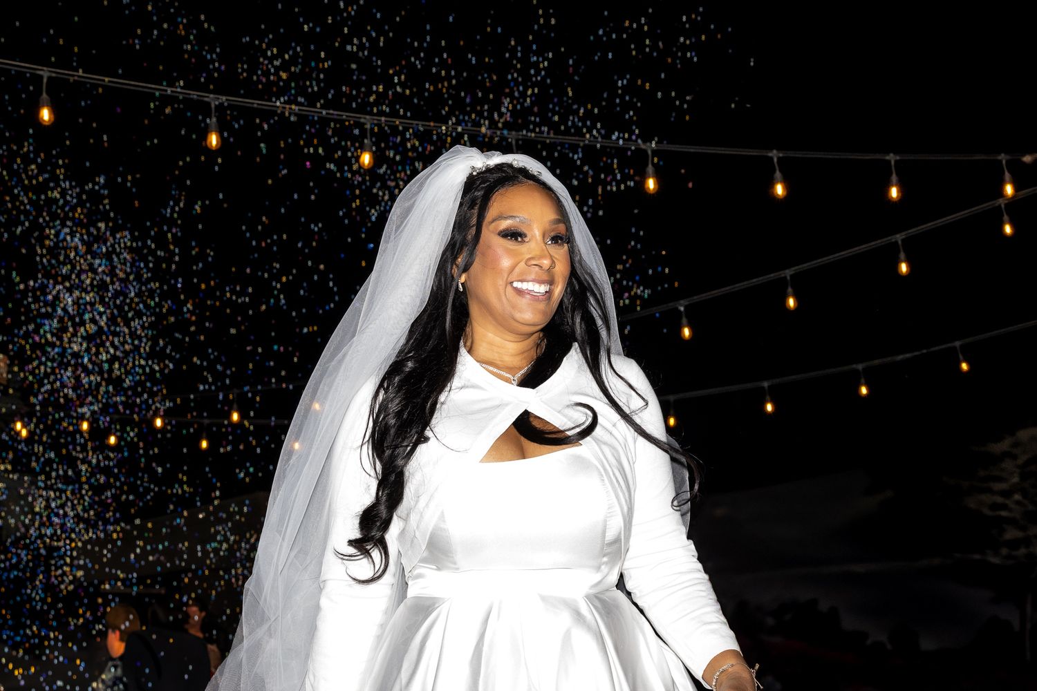 Smiling bride in white satin gown and veil under sparkling string lights at night wedding.