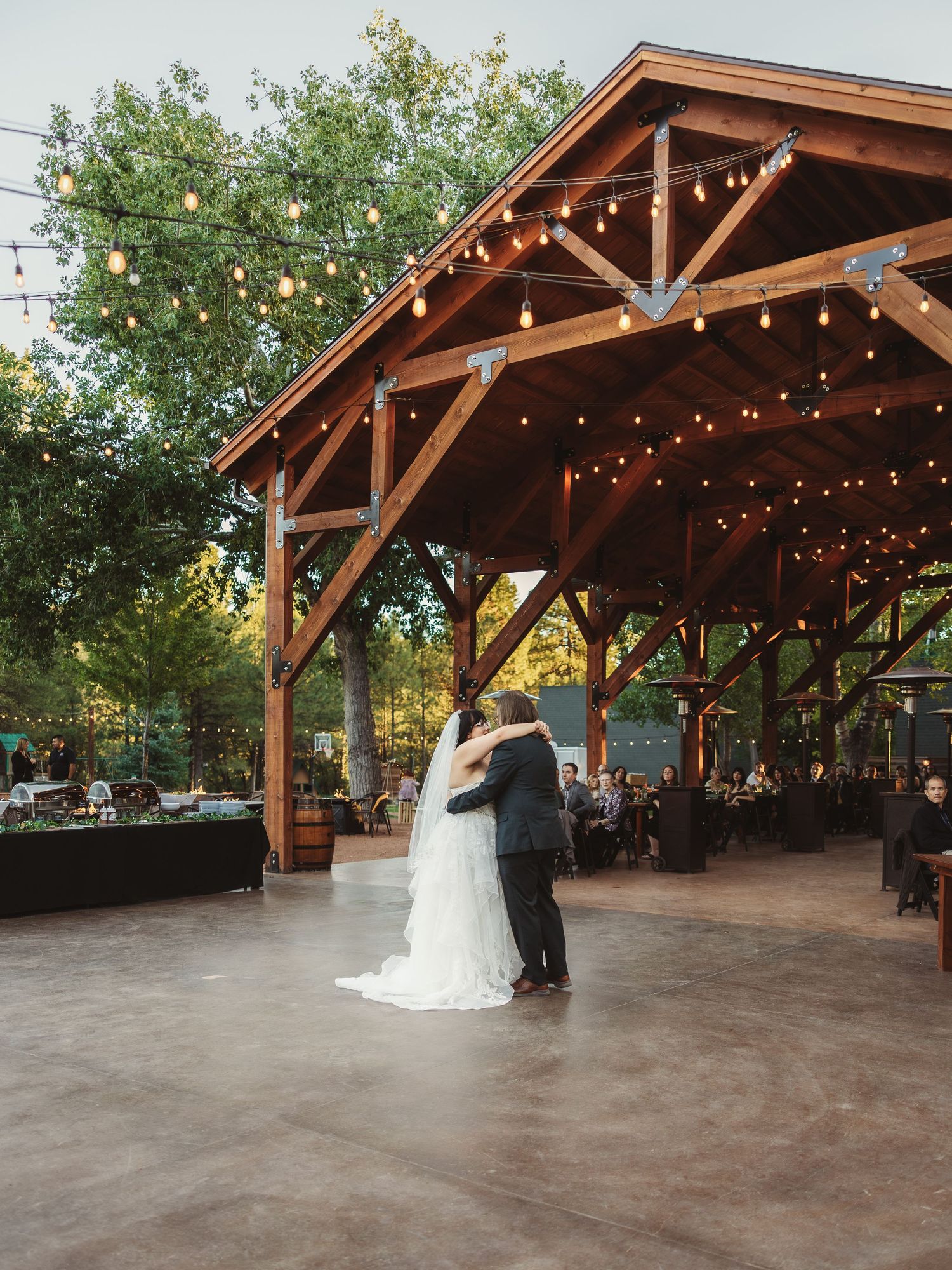 Bride and groom sharing first dance under rustic wooden pavilion with string lights at outdoor reception.