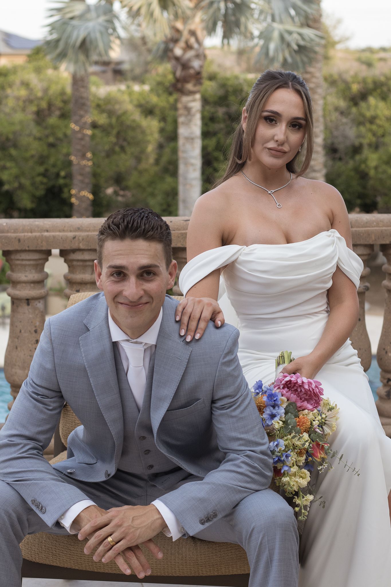 Smiling bride and groom posing at outdoor wedding venue, bride holding colorful wildflower bouquet.