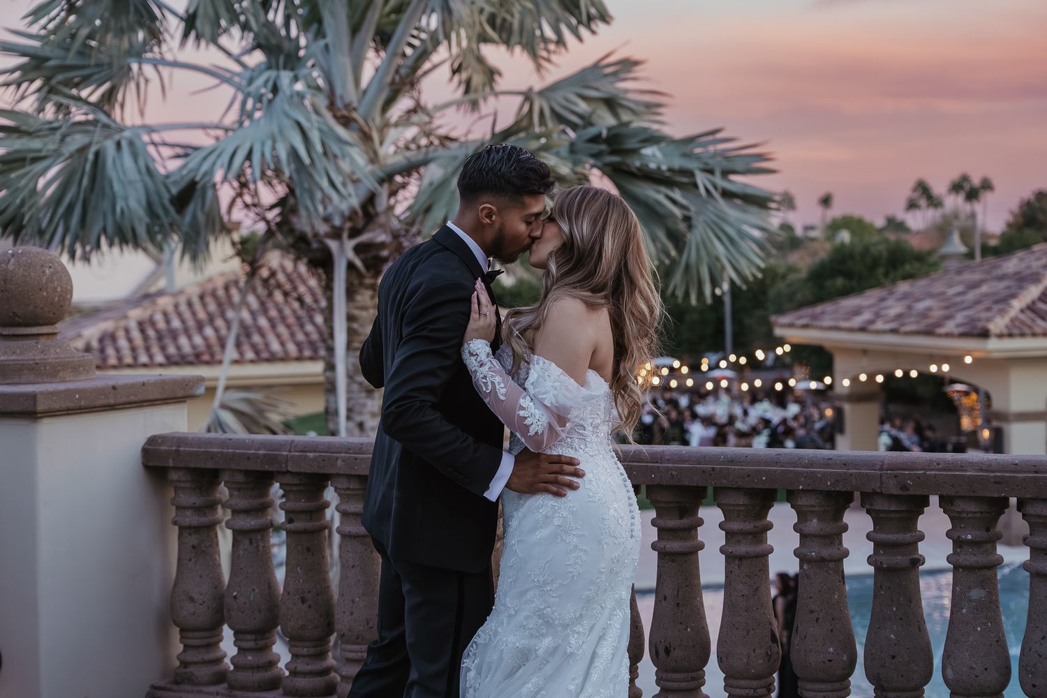 Newlyweds share a passionate kiss on an elegant balcony during a beautiful pink and purple sunset.