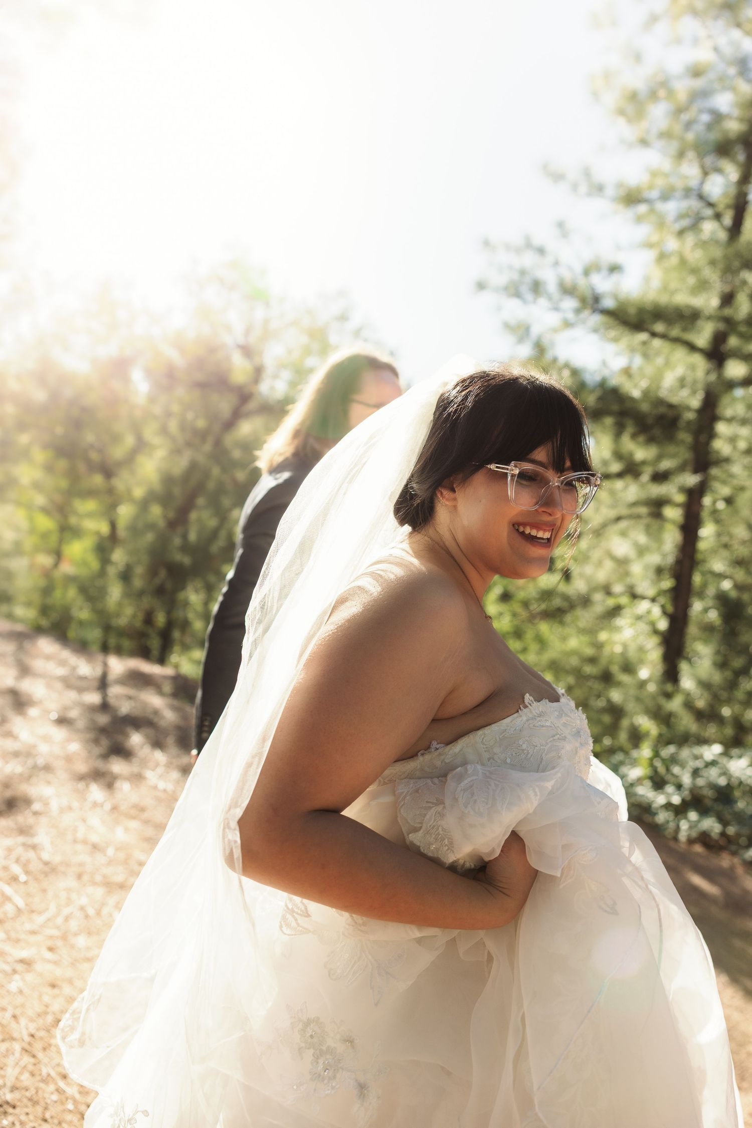 Smiling bride in white gown and veil with glasses looks back during outdoor forest wedding photo session.