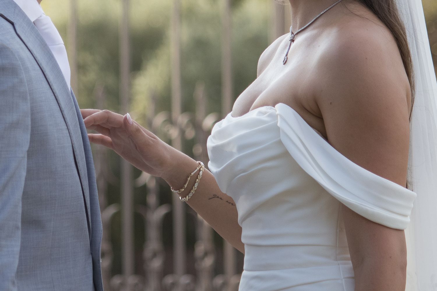 Bride in elegant off-shoulder white wedding gown standing beside groom in light gray suit outdoors.
