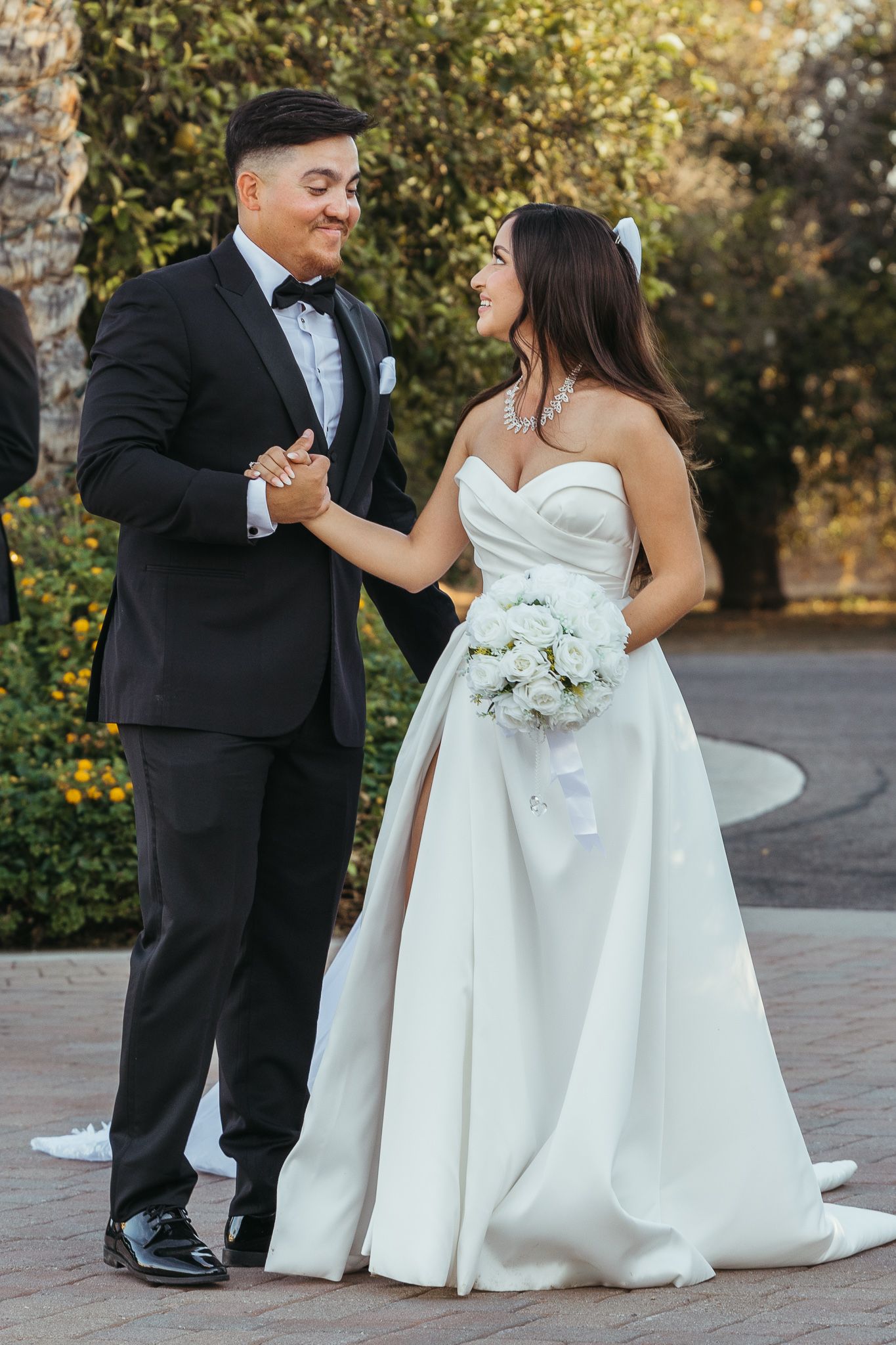 Smiling groom in black tuxedo holds hands with bride in strapless ballgown outdoors.