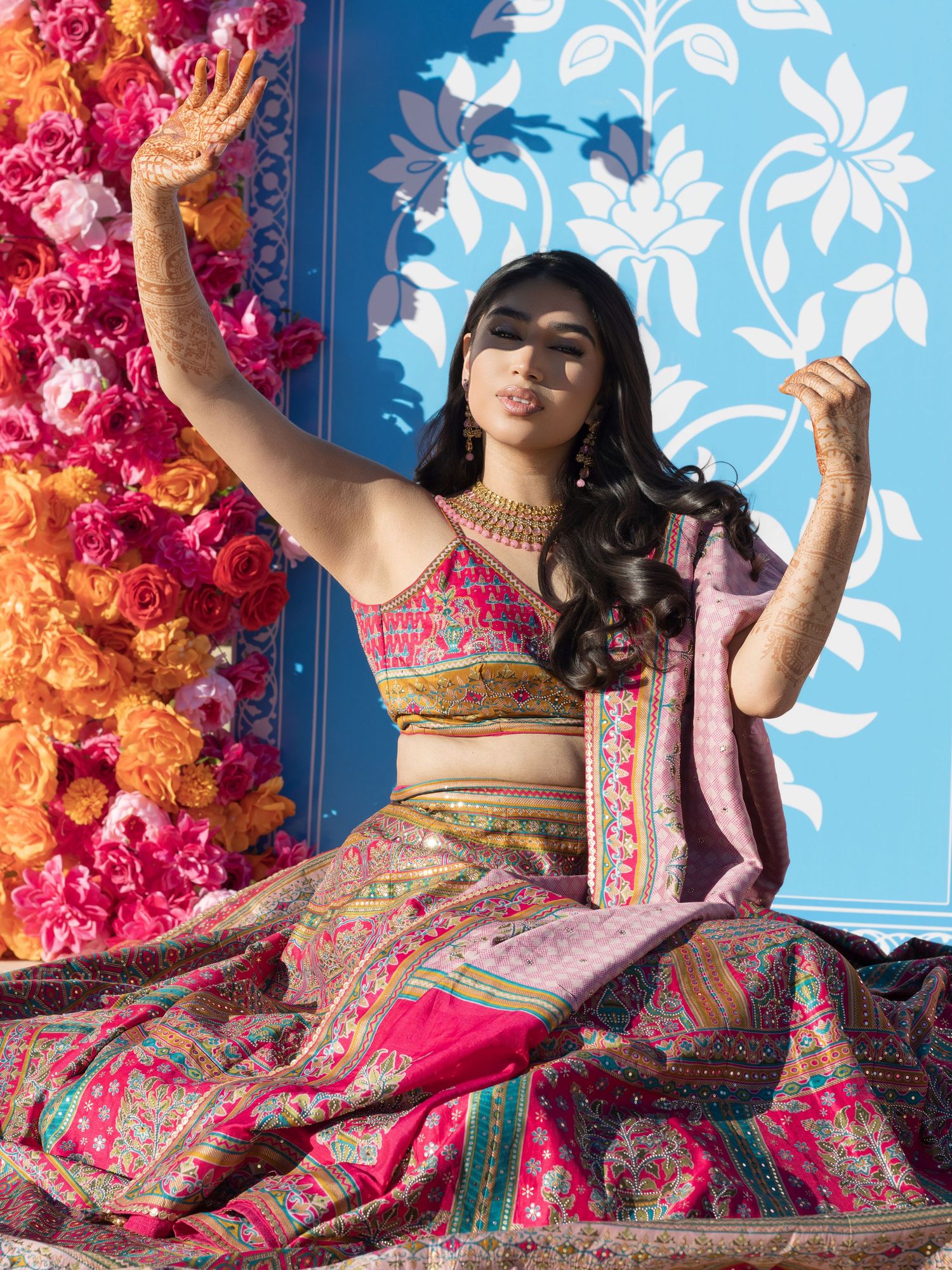 Woman in vibrant pink lehenga with henna hands posed against floral backdrop at Indian wedding celebration.