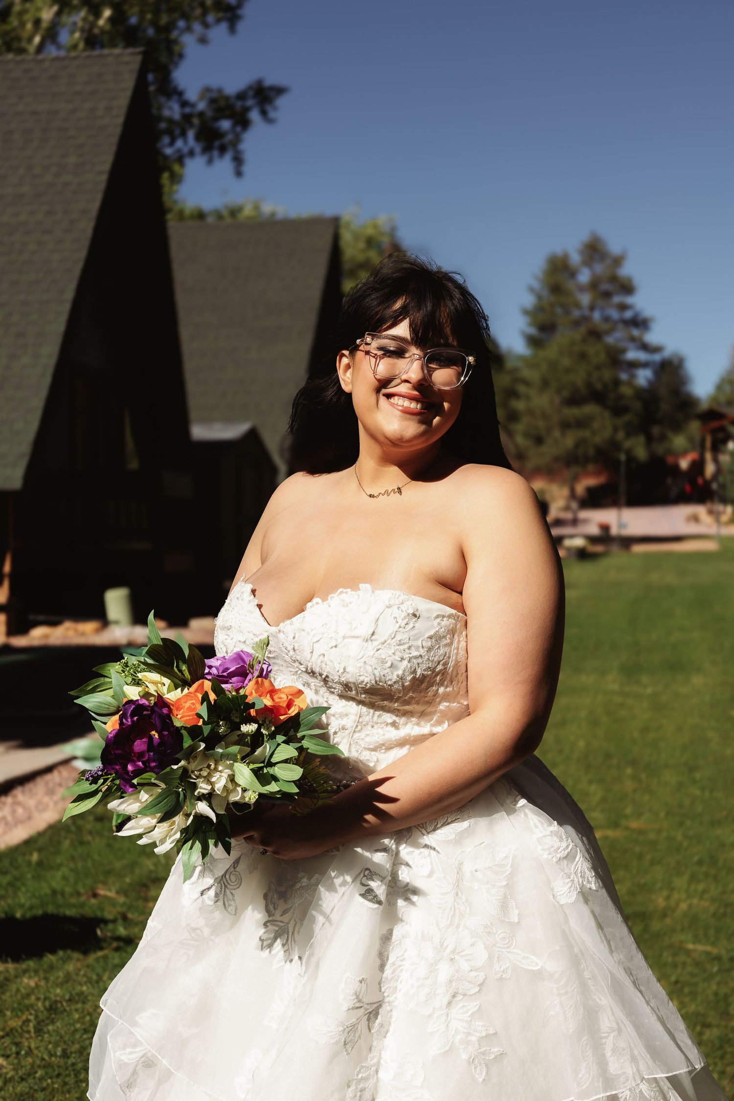 Smiling bride in strapless lace gown holds colorful bouquet outdoors near rustic cabin venue.