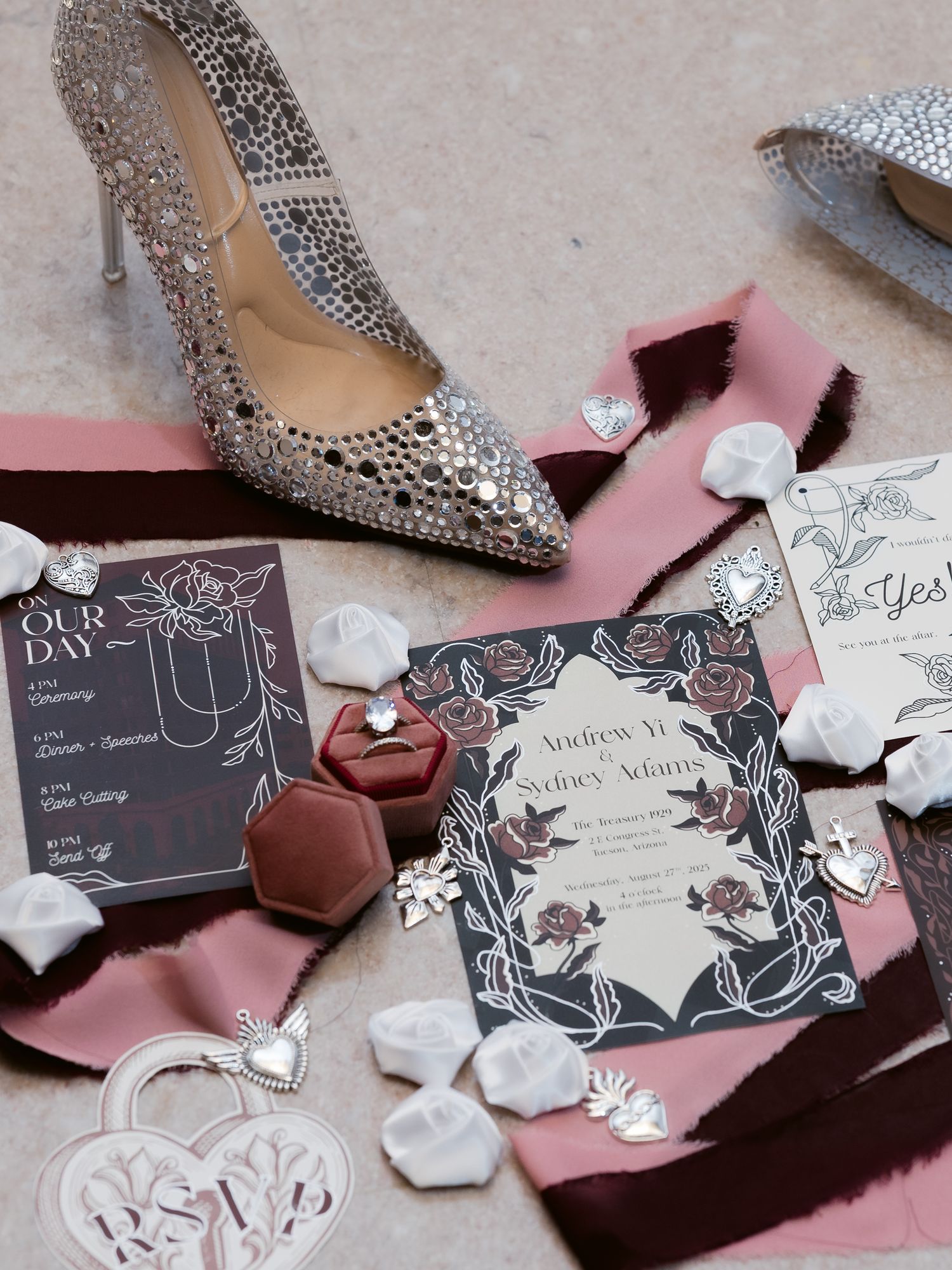 Wedding flat lay featuring crystal heels, floral invitations, velvet ring box, and pearl accessories on pink ribbons.