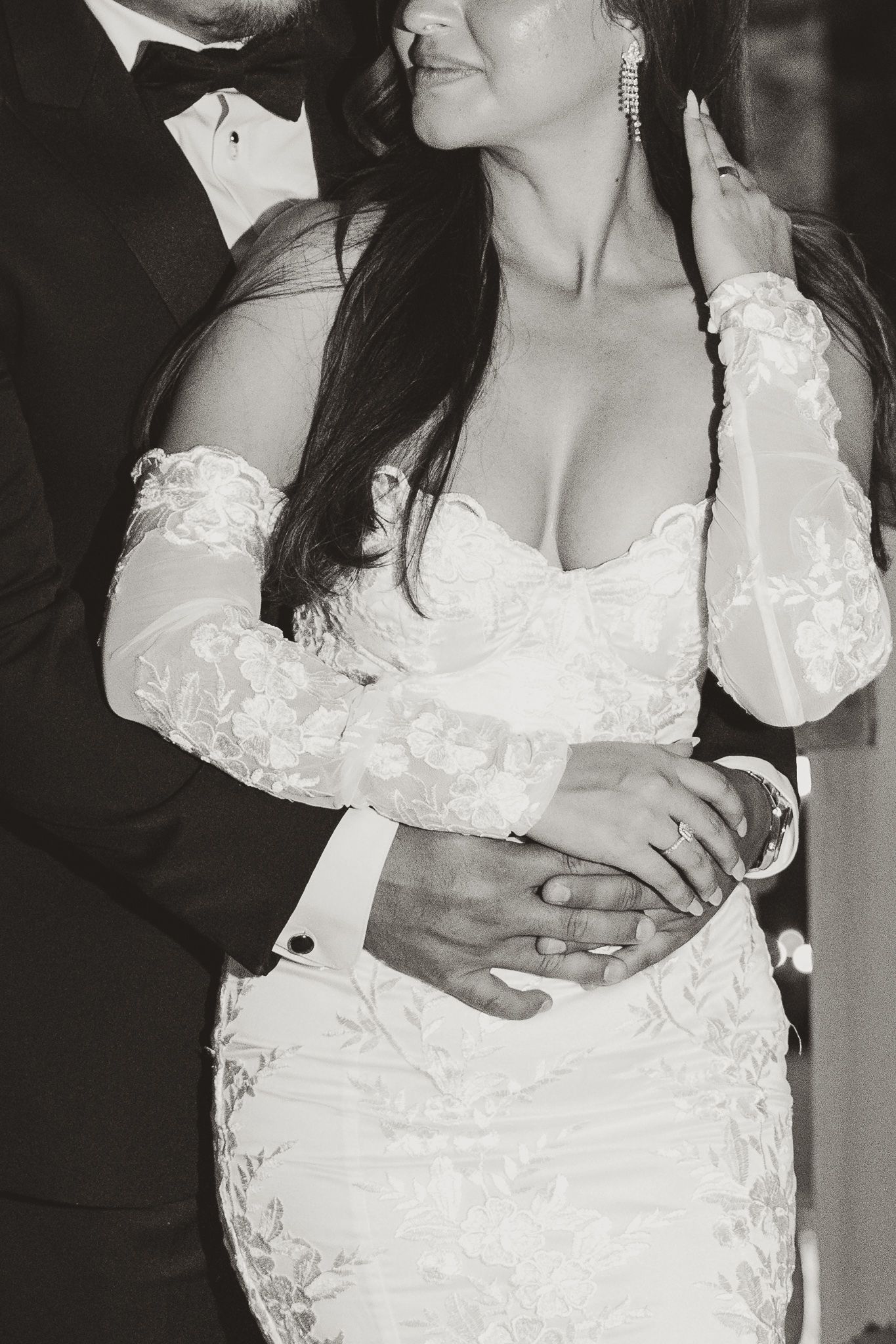 Black and white photo of groom embracing bride in lace off-shoulder wedding gown, hands clasped.