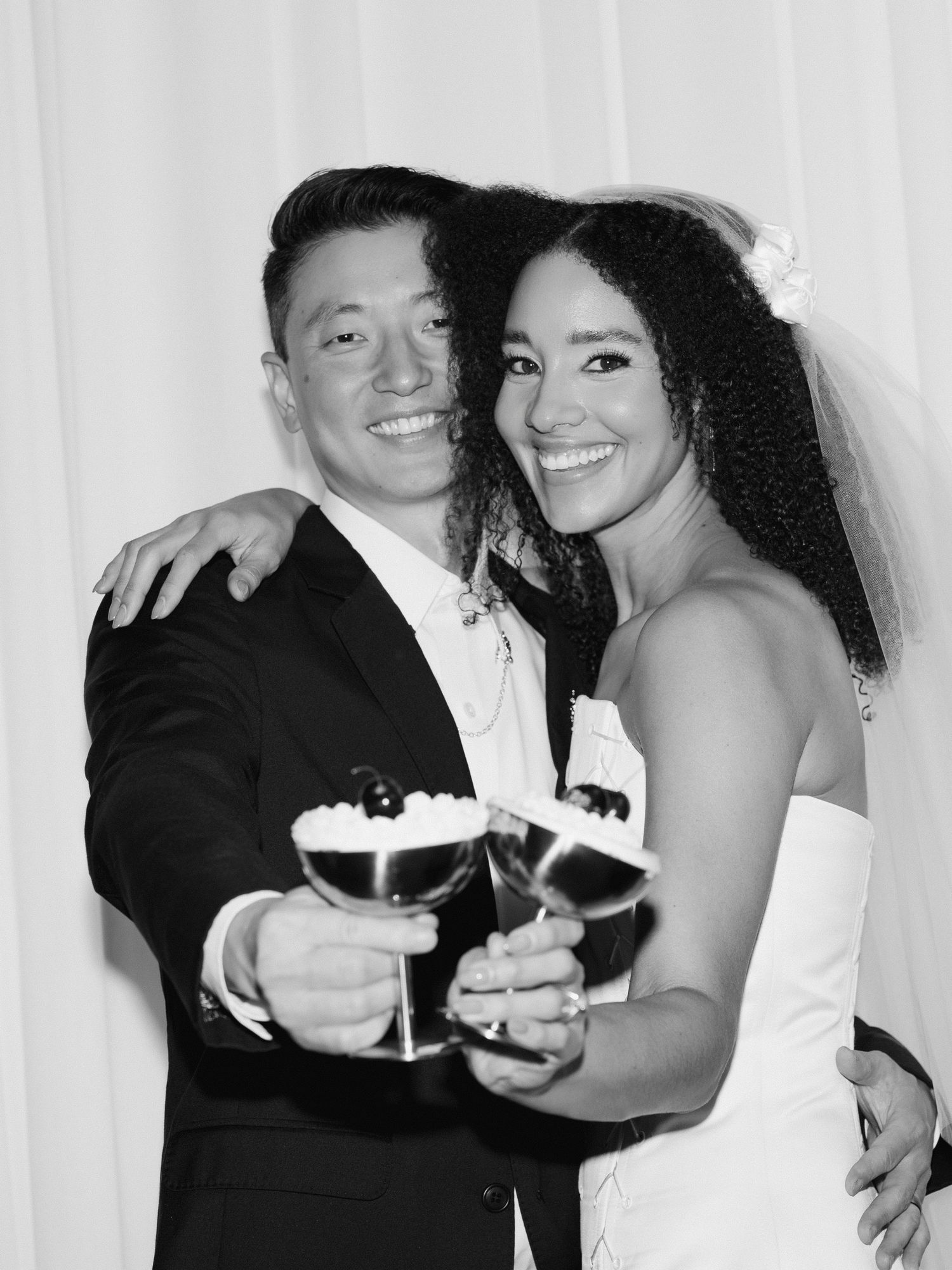 Black and white photo of smiling newlyweds toasting with cocktails at their wedding reception.