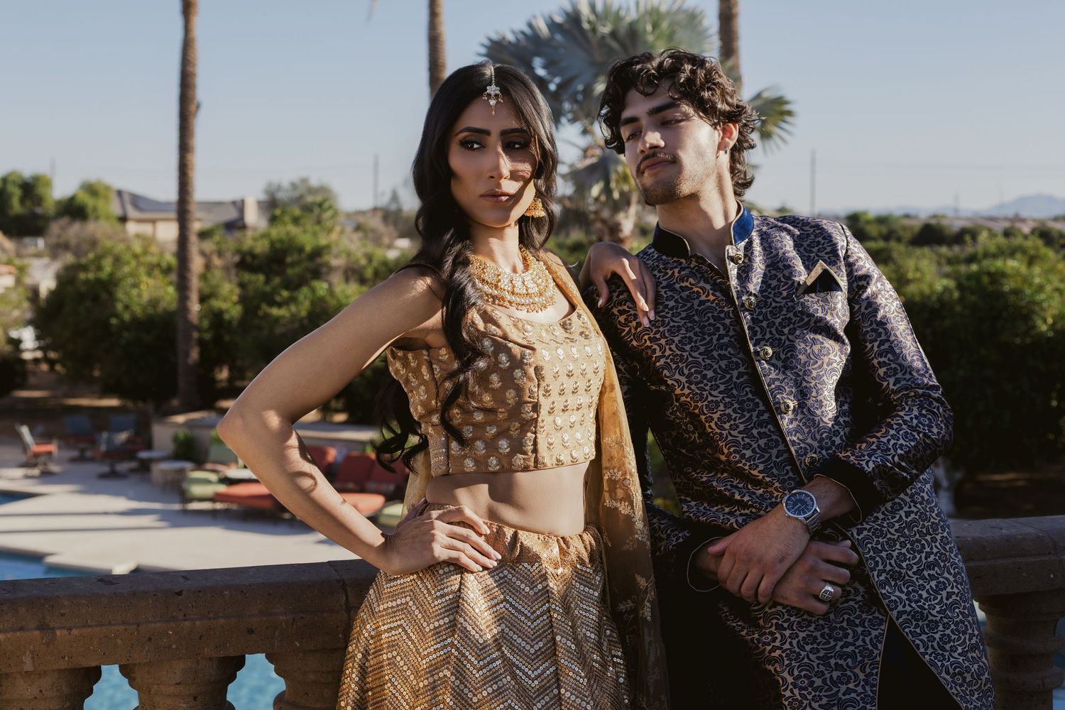 Stylish South Asian couple in colorful Indian fusion outfits posing outdoors at a sunny estate.