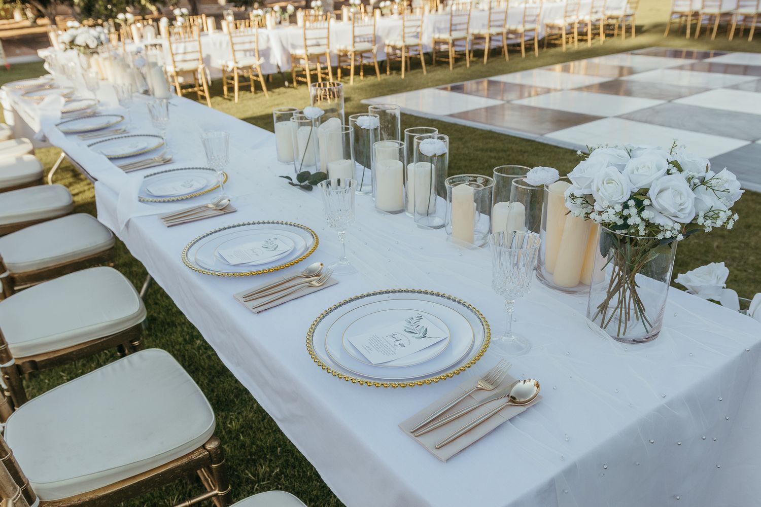 Outdoor wedding reception table with white linen, gold-rimmed plates, candles, and white floral centerpiece.