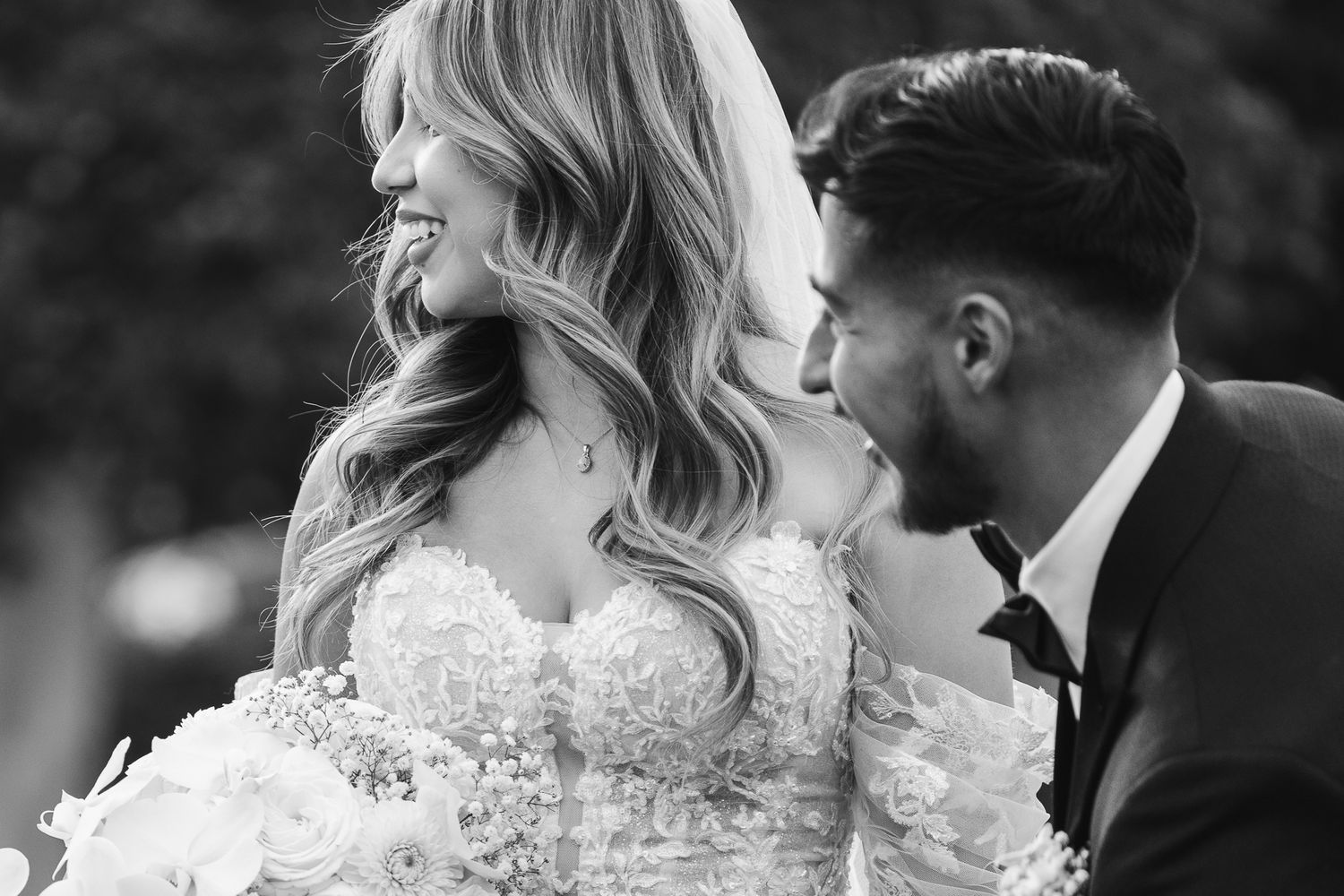 Black and white photo of groom whispering to laughing bride holding a white floral bouquet.