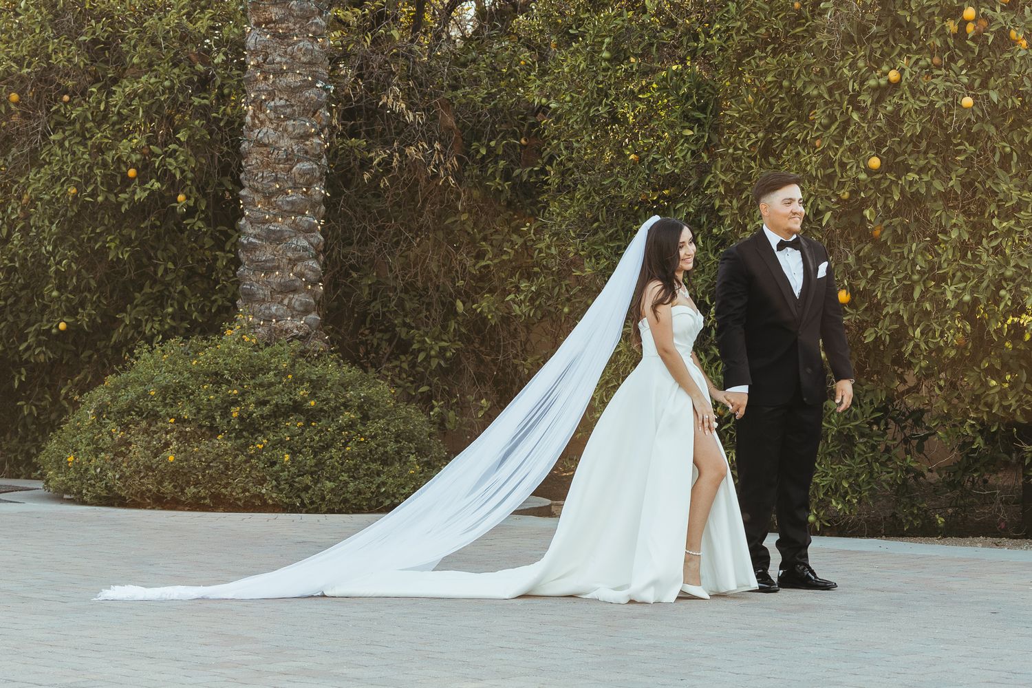 Bride in flowing white gown with long veil stands beside tuxedo-clad groom outdoors.