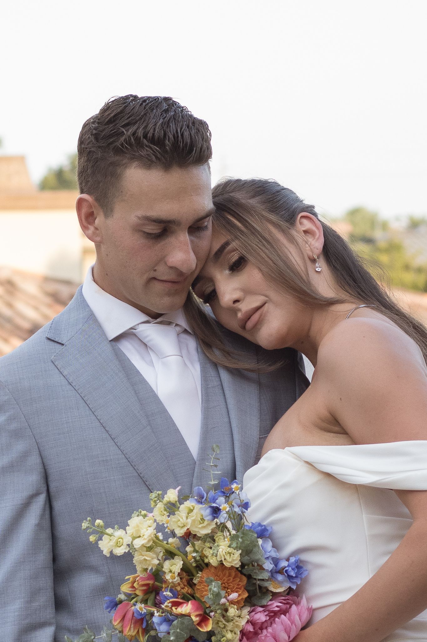 Romantic bride and groom portrait, bride resting head on grooms shoulder holding colorful bouquet.