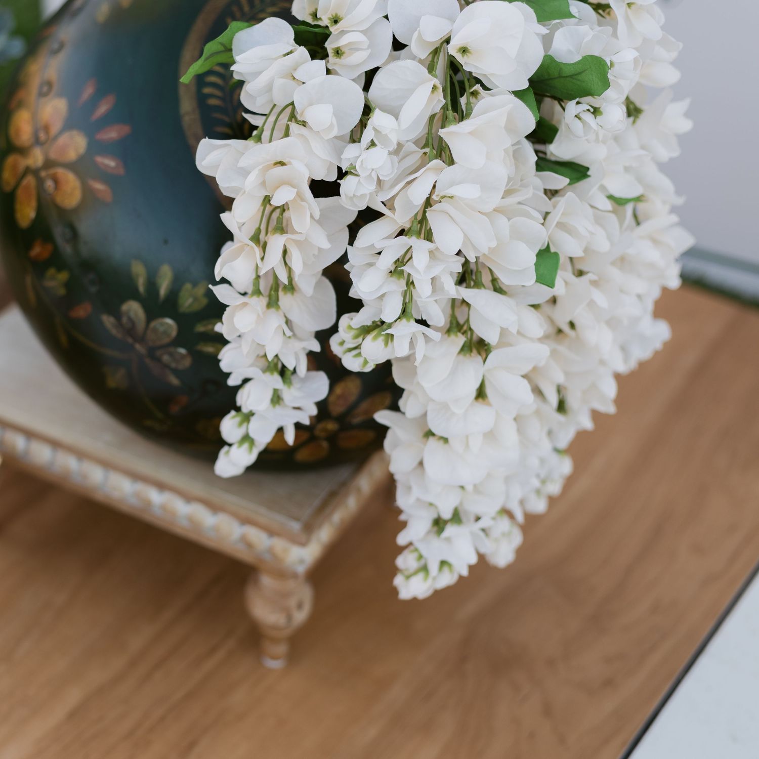 White wisteria flowers cascade from a decorative painted vase on a wooden stand.
