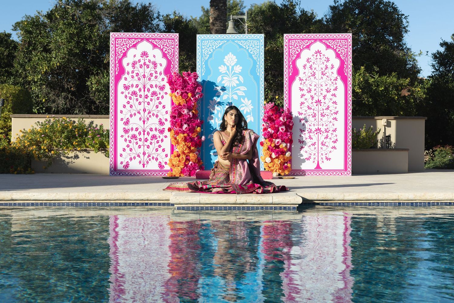 Woman in colorful Indian outfit seated poolside before vibrant pink and blue Mughal-inspired panels.