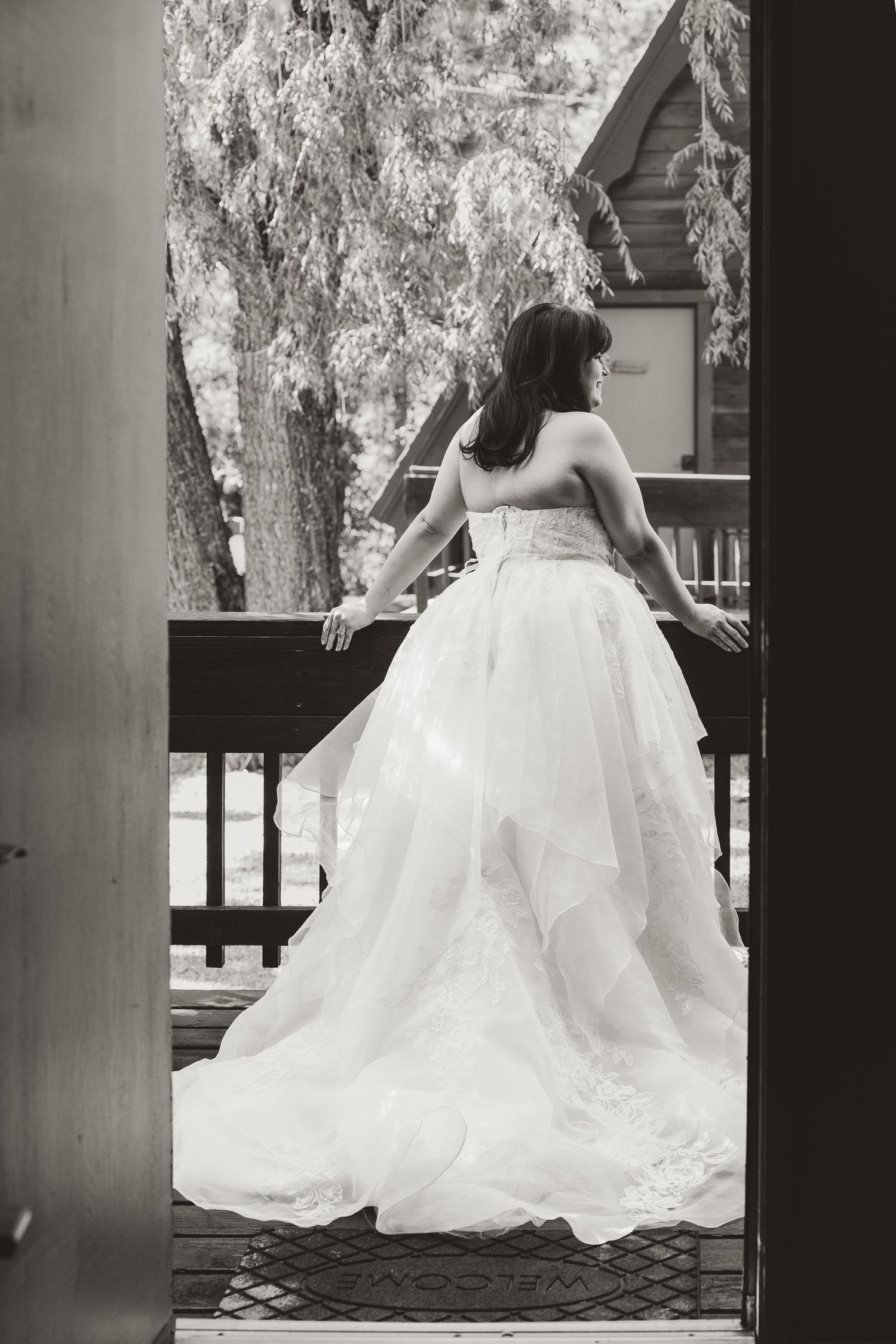 Bride in voluminous strapless ballgown with lace detail stands on wooden deck in black and white photo.