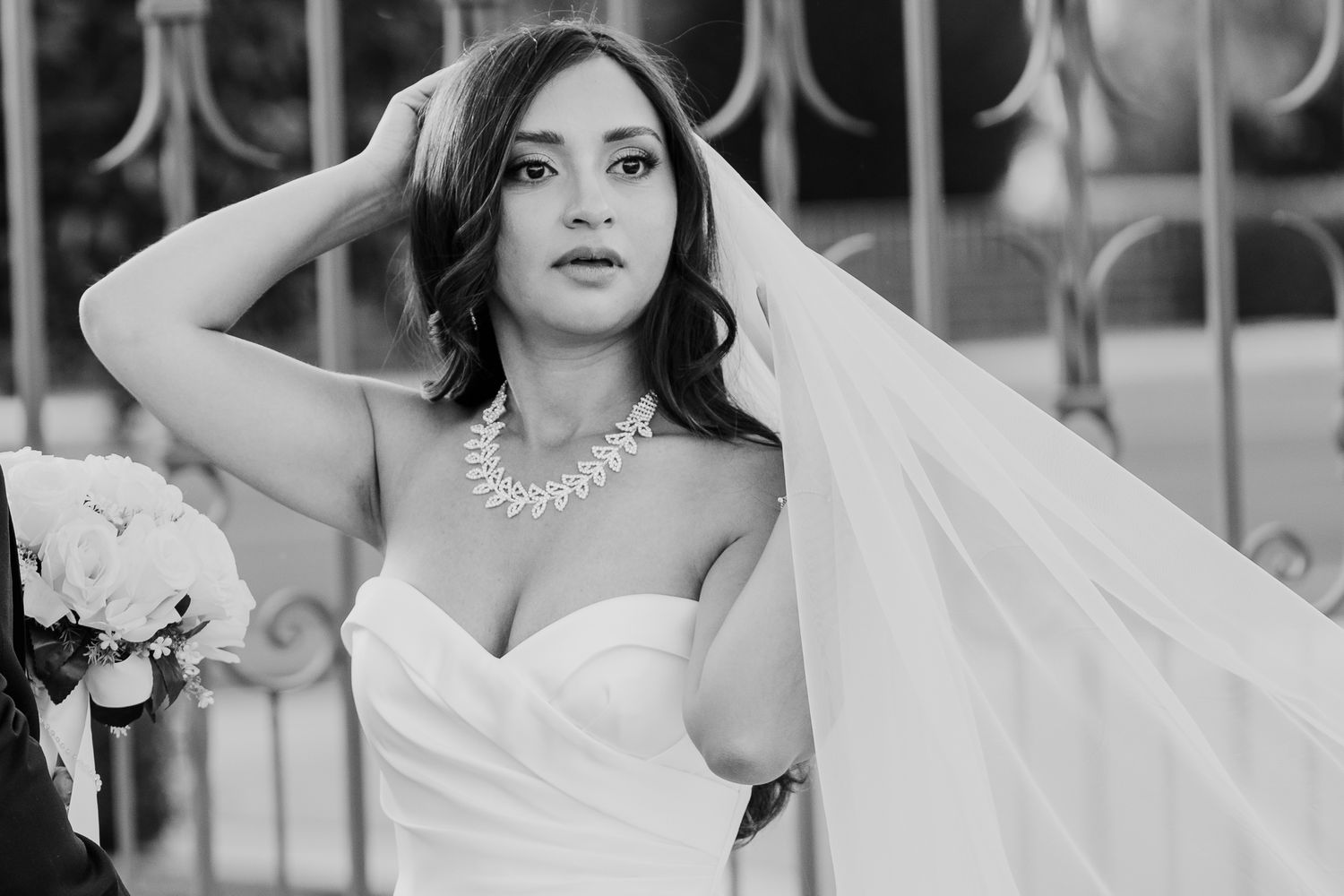 Black and white portrait of bride in strapless gown adjusting veil with pearl necklace.