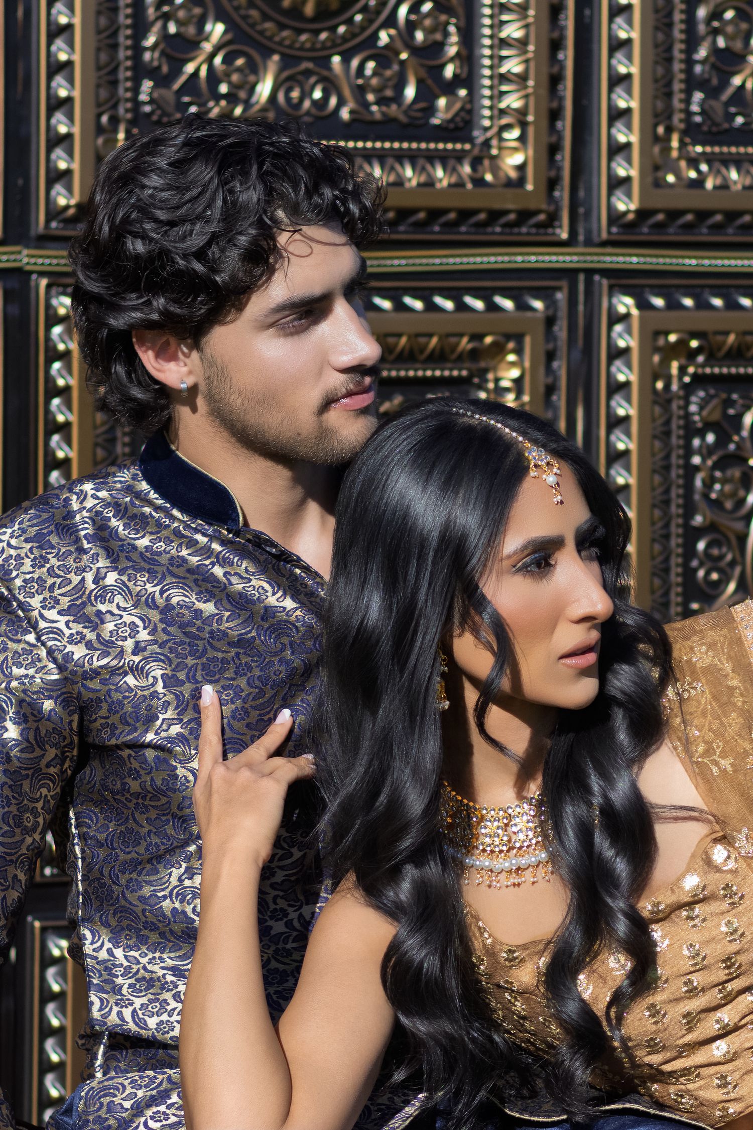 Elegant couple in traditional Indian wedding attire posing before ornate golden decorative door backdrop.