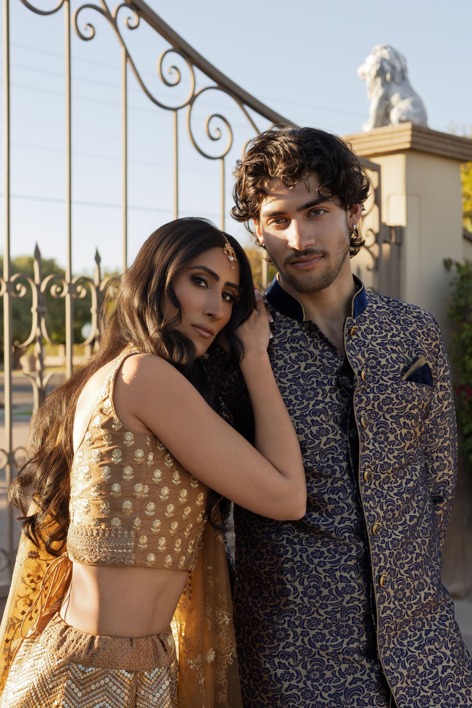 Couple in elegant Indian ethnic wear posing intimately near decorative iron gates outdoors.