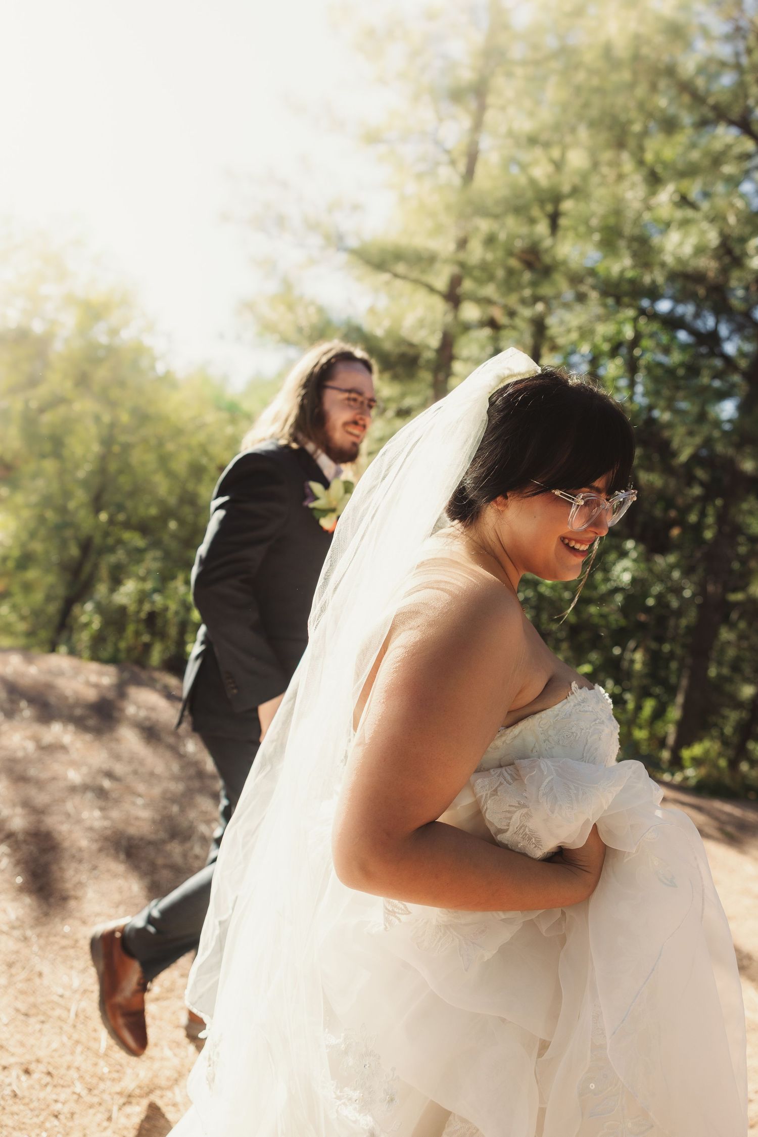 Bride in white gown and veil smiles back at groom in black suit during outdoor wedding photo.