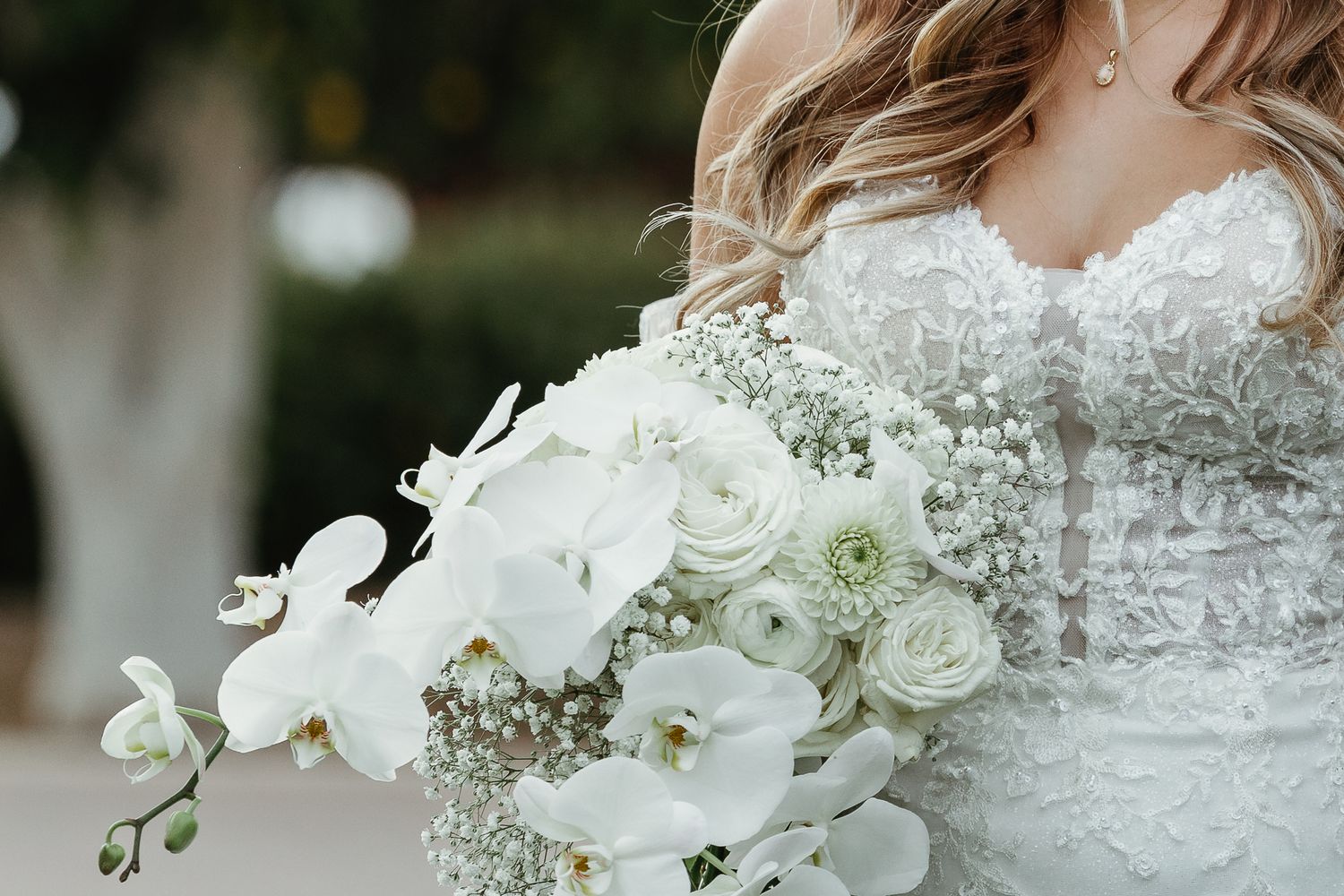 Close-up of bride holding a stunning white bouquet of orchids, roses, and baby's breath against a lace wedding gown.