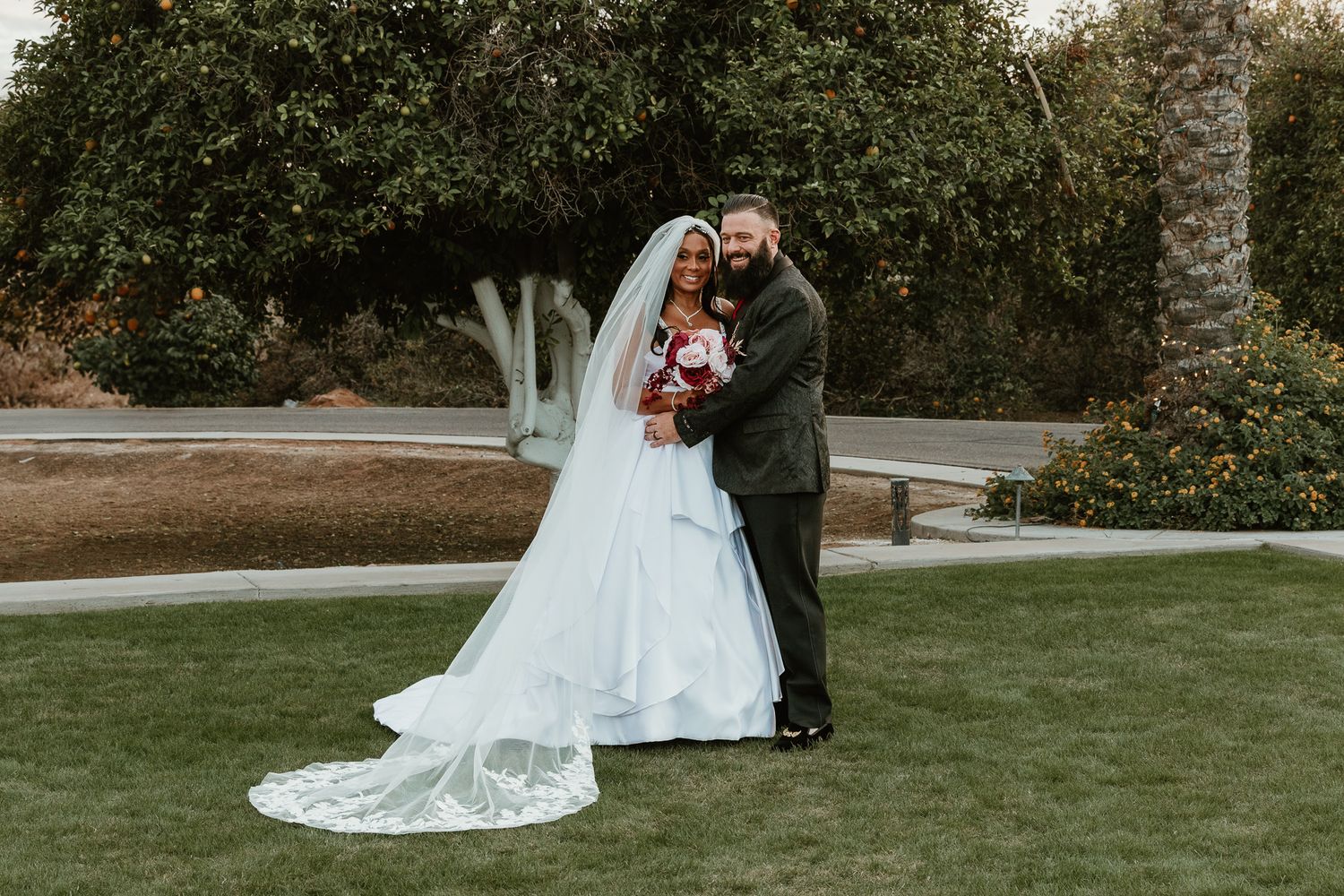 Bride and groom posing together on green lawn, bride in white gown with long cathedral veil.