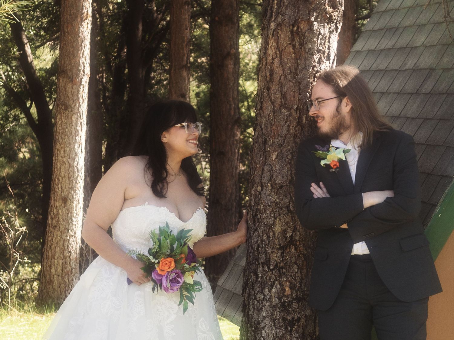 Bride and groom share a romantic moment leaning against pine trees in a forest wedding setting.