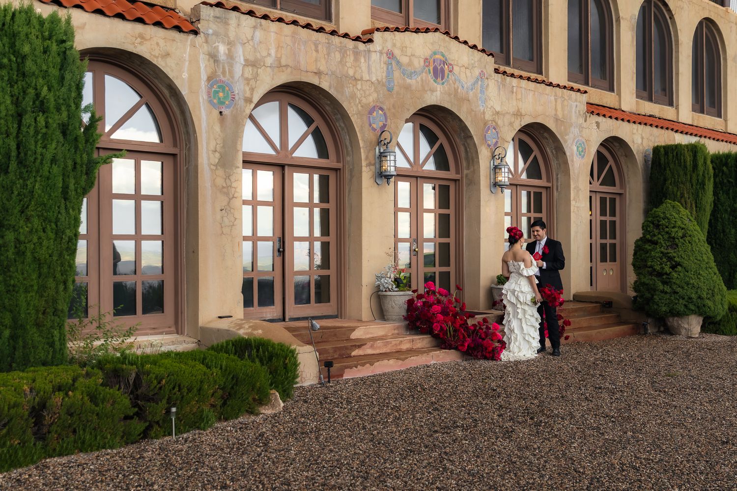 Bride and groom pose outside an elegant Spanish-style wedding venue with arched windows and red floral decor.