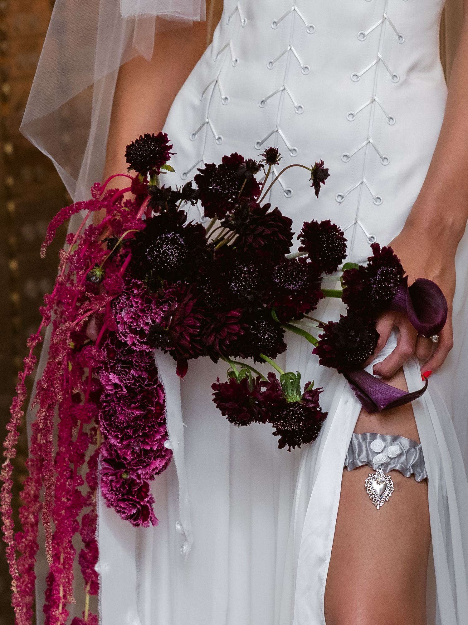 Bride holding dramatic dark burgundy and magenta cascading wedding bouquet with scabiosa blooms and silver heart garter.