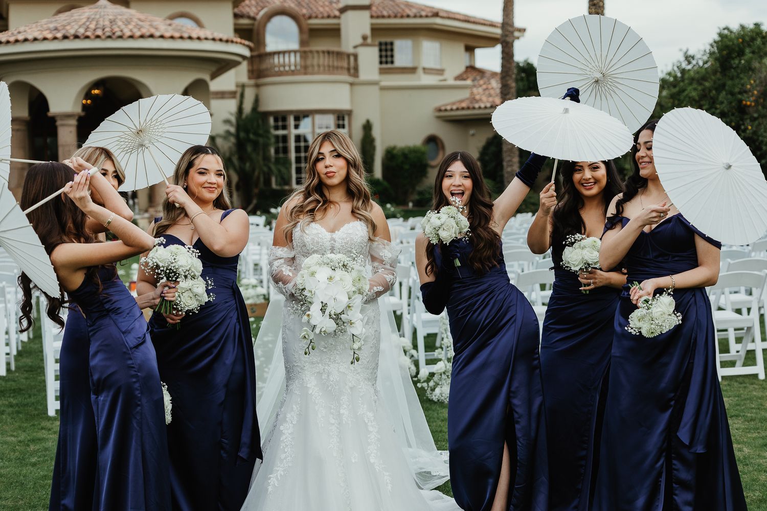 Bride with bridesmaids in navy blue dresses holding white parasols and bouquets at outdoor wedding ceremony.