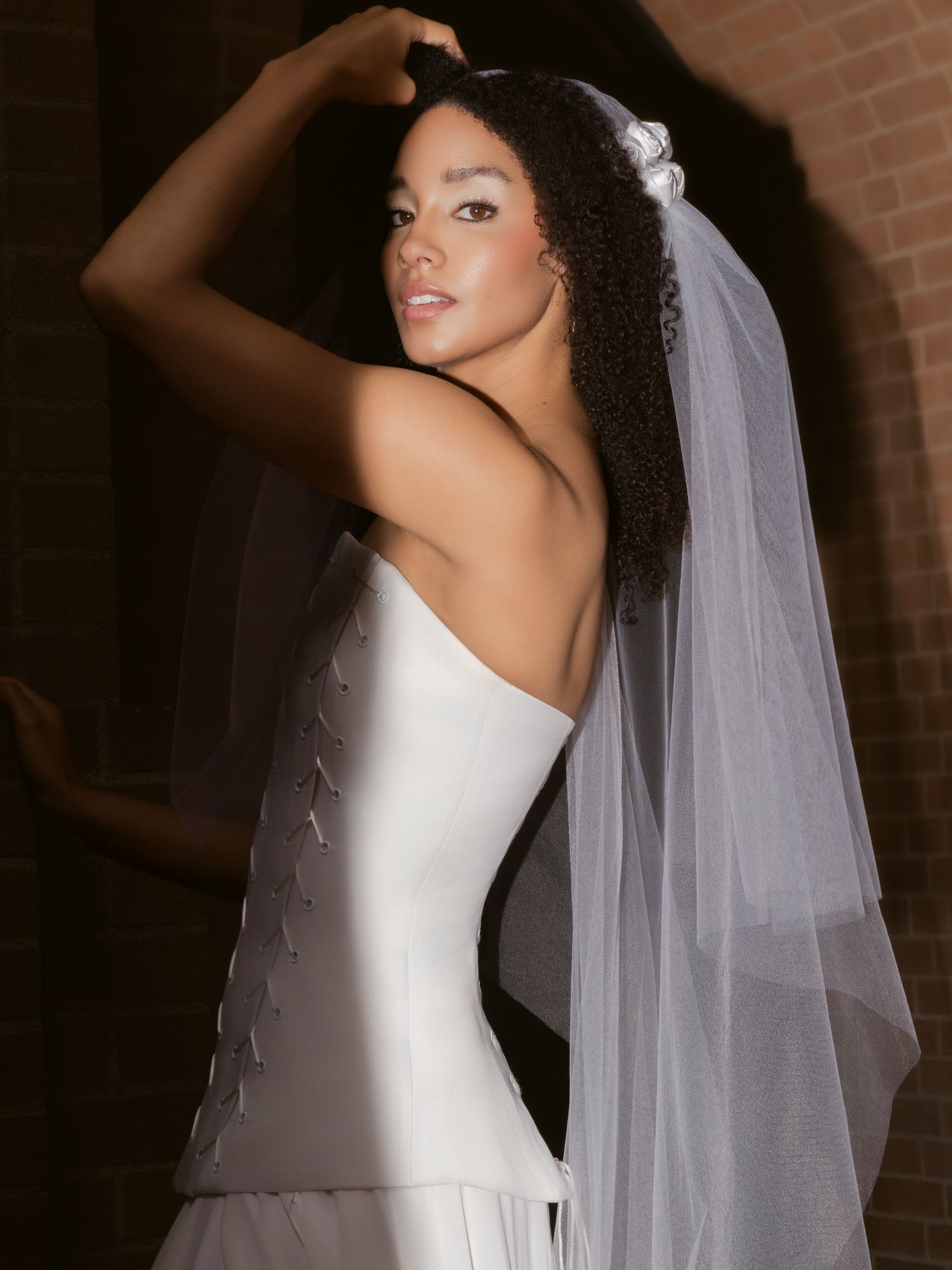 Bride in strapless white gown with lace back detail and flowing veil, posing elegantly against brick wall.