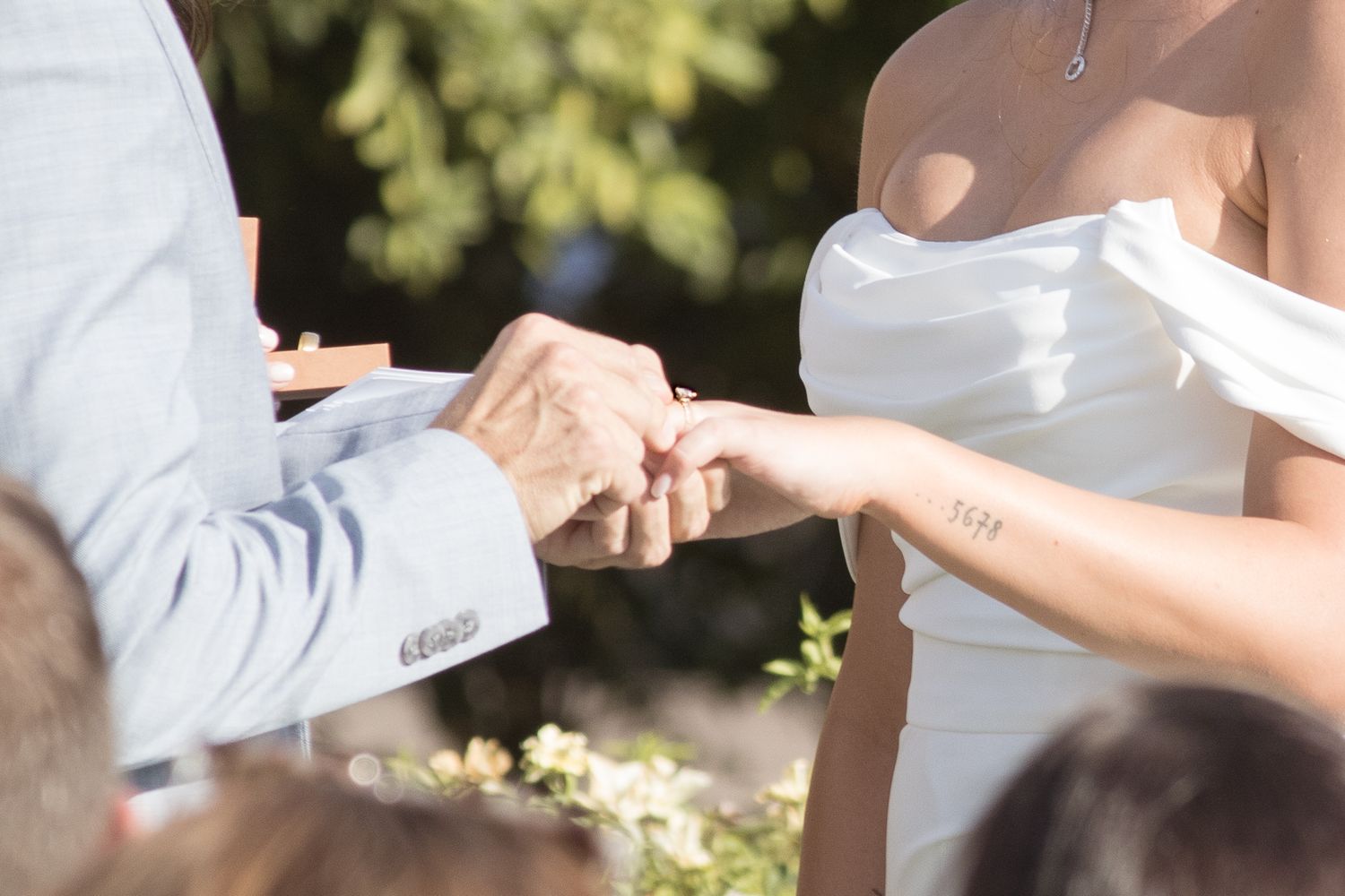 Bride in white off-shoulder gown exchanging rings with groom during outdoor wedding ceremony.