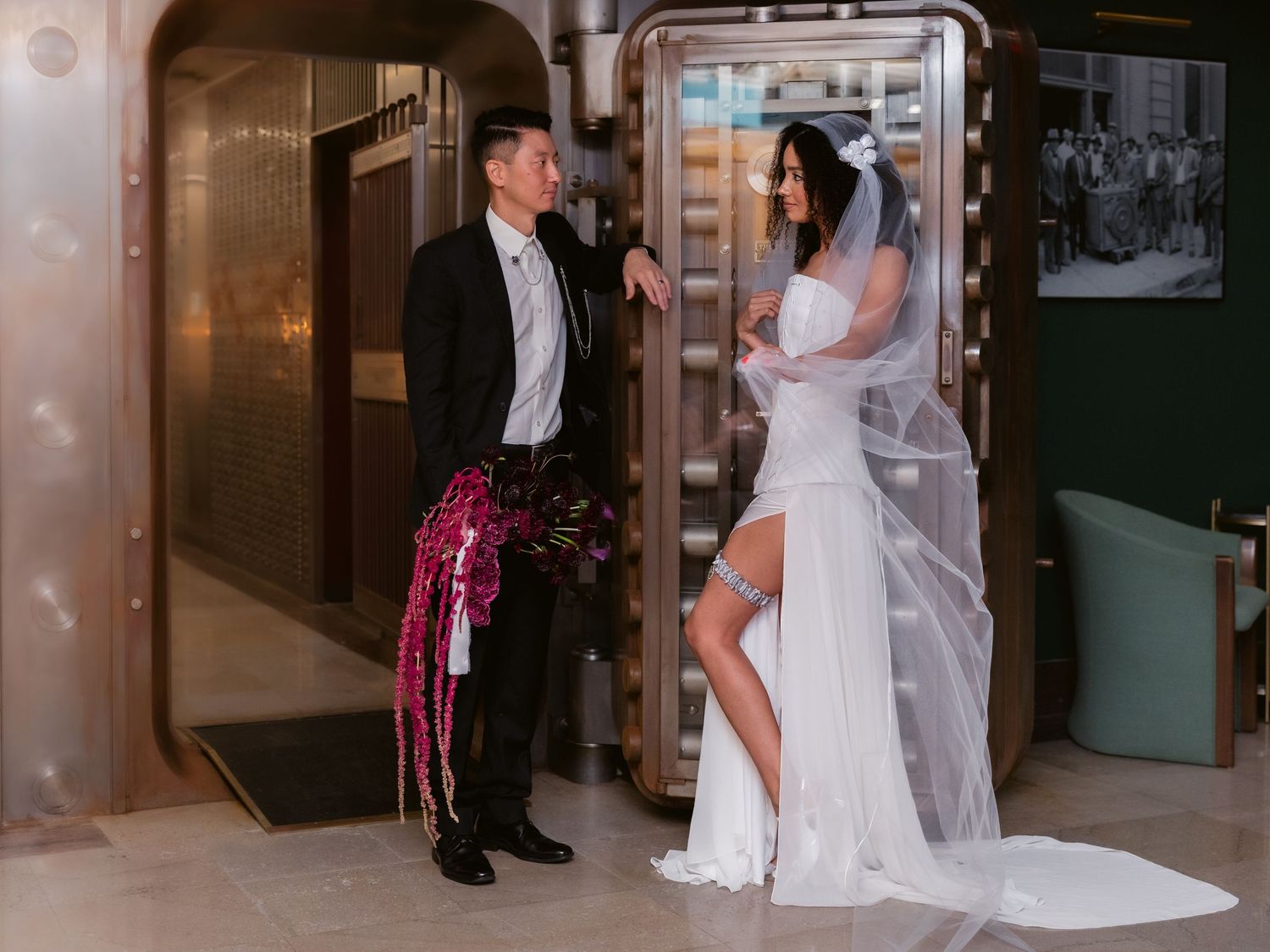 Bride and groom standing at ornate vault door, bride in fitted white gown with thigh slit holding pink floral streamer.