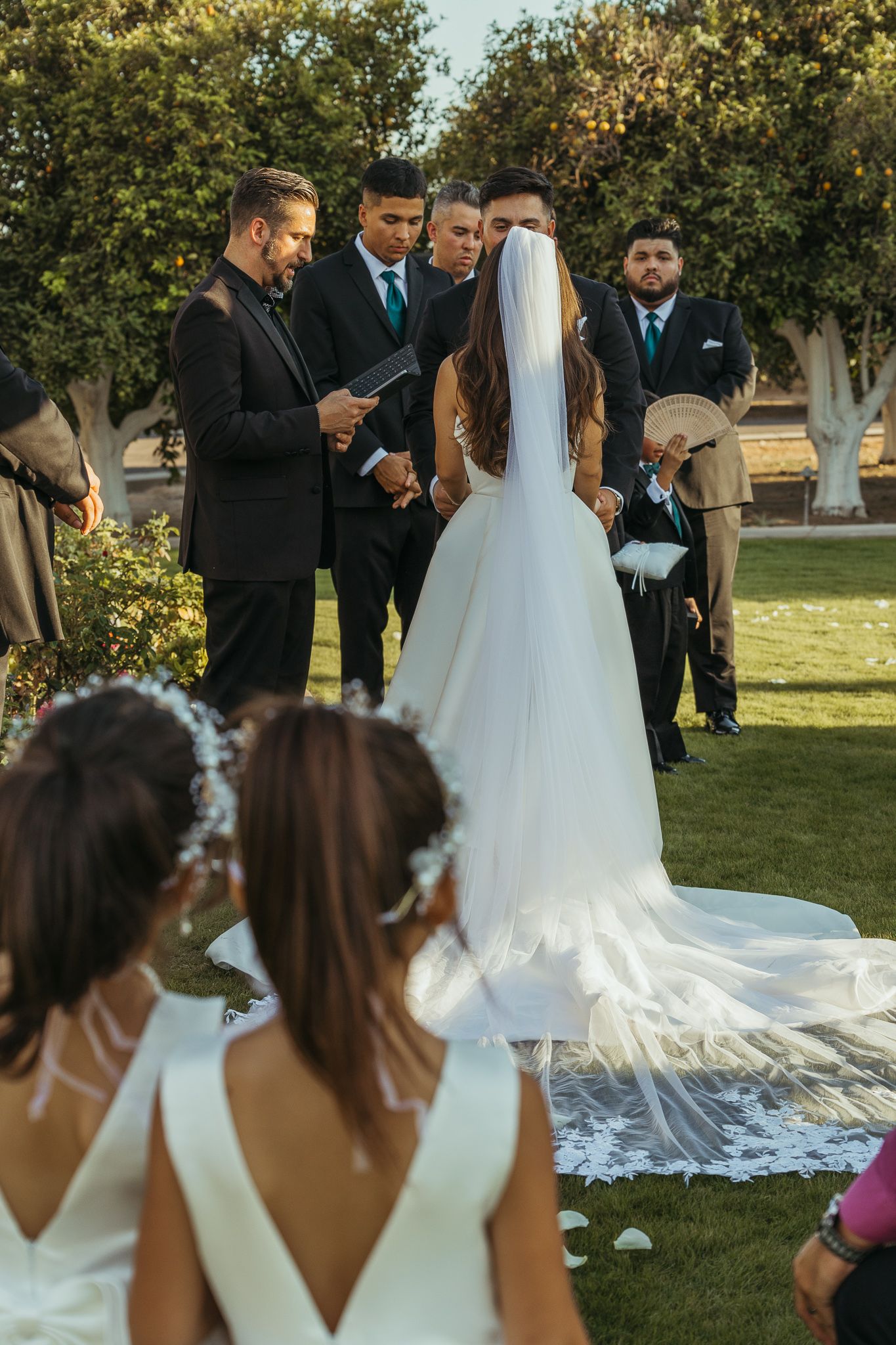 Bride in lace-trimmed cathedral veil walks toward groom and groomsmen at outdoor ceremony.